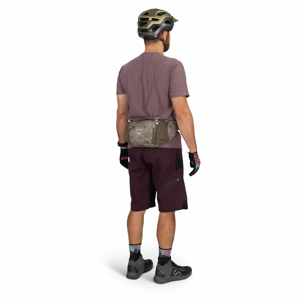 The image depicts a person from behind wearing biking gear. The individual has a gray and olive green Osprey waist pack around their waist, complementing their biking outfit. They are dressed in a maroon t-shirt and matching maroon shorts with a side pock