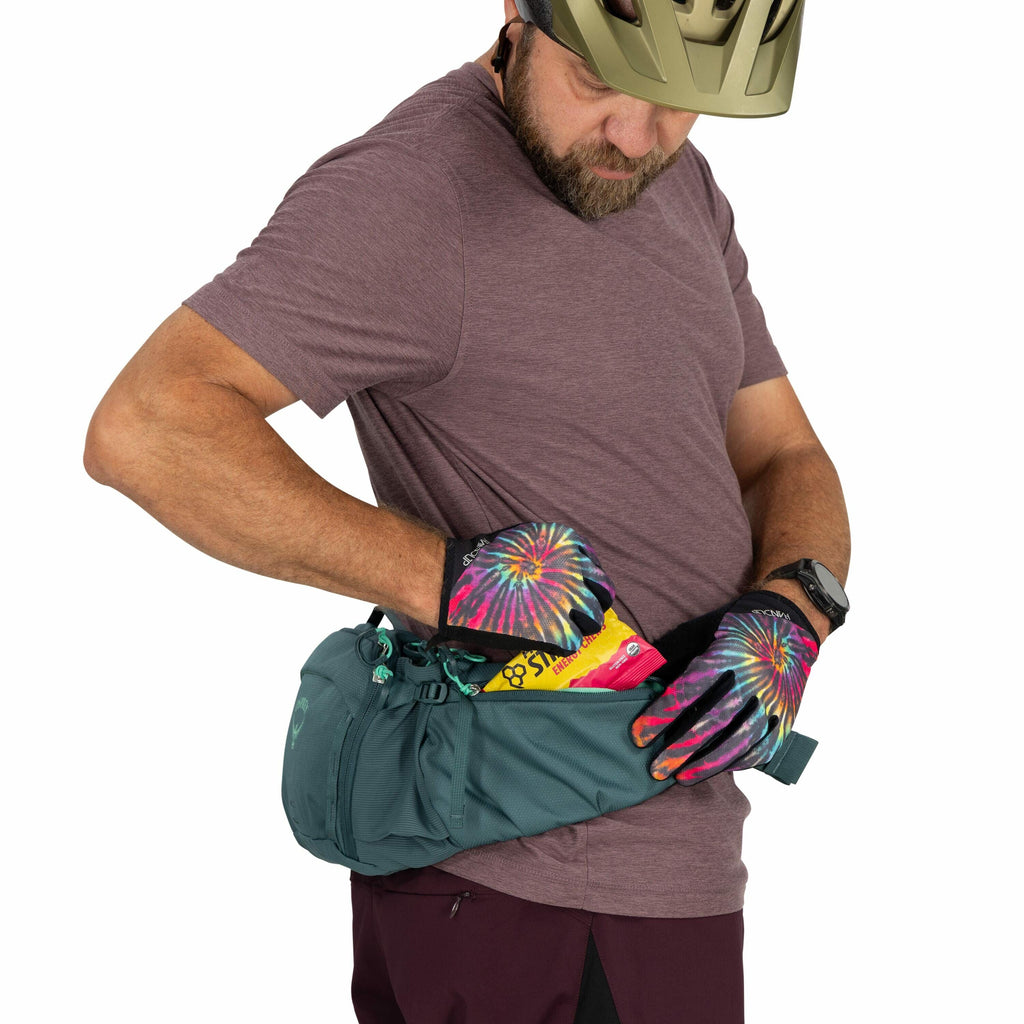 The image shows a person wearing a maroon t-shirt and dark pants, accessorized with a wristwatch and a helmet. They are wearing gloves with a vibrant tie-dye pattern in pink, blue, yellow, and green. The person is adjusting or retrieving items from a dark