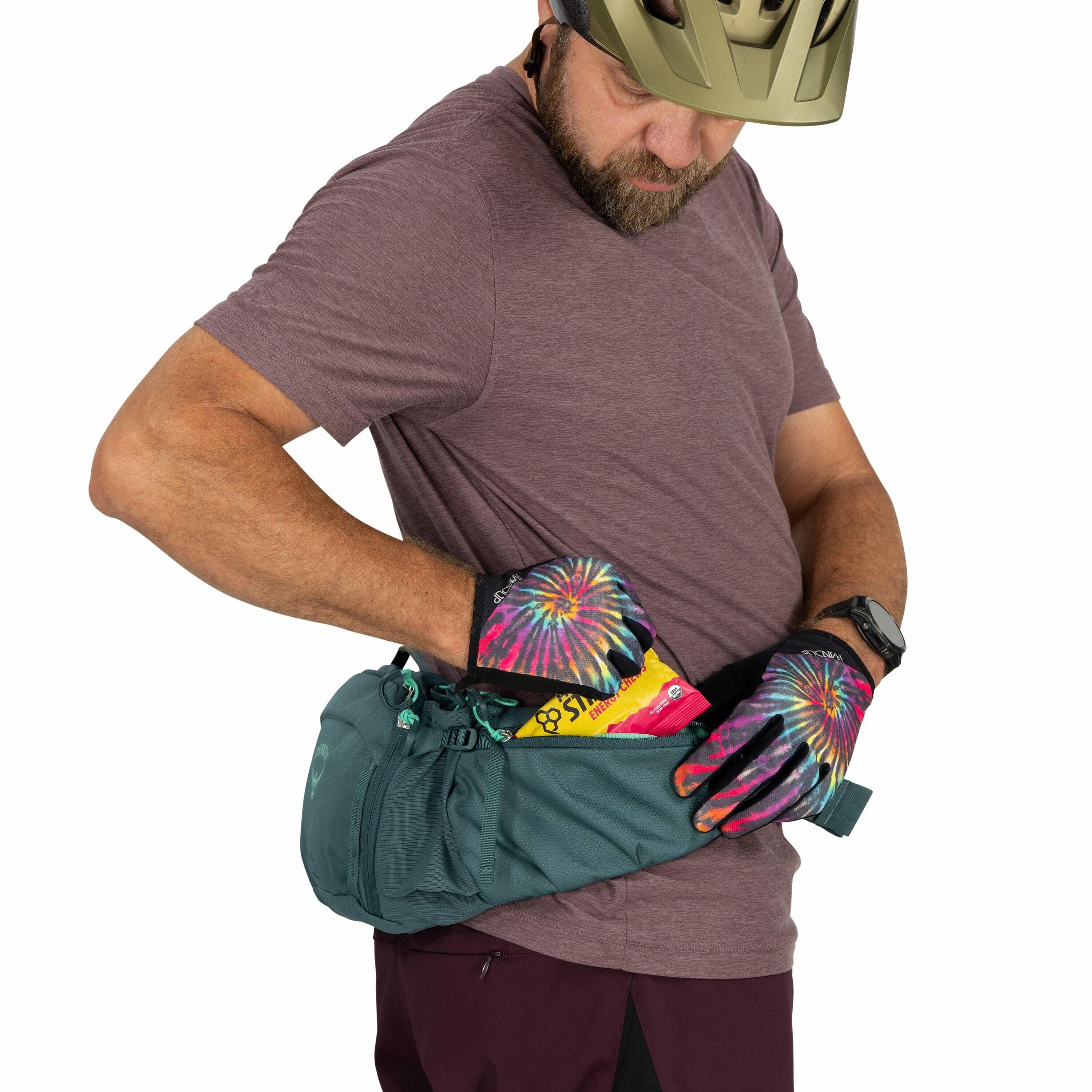 The image shows a person wearing a maroon t-shirt and dark pants, accessorized with a wristwatch and a helmet. They are wearing gloves with a vibrant tie-dye pattern in pink, blue, yellow, and green. The person is adjusting or retrieving items from a dark