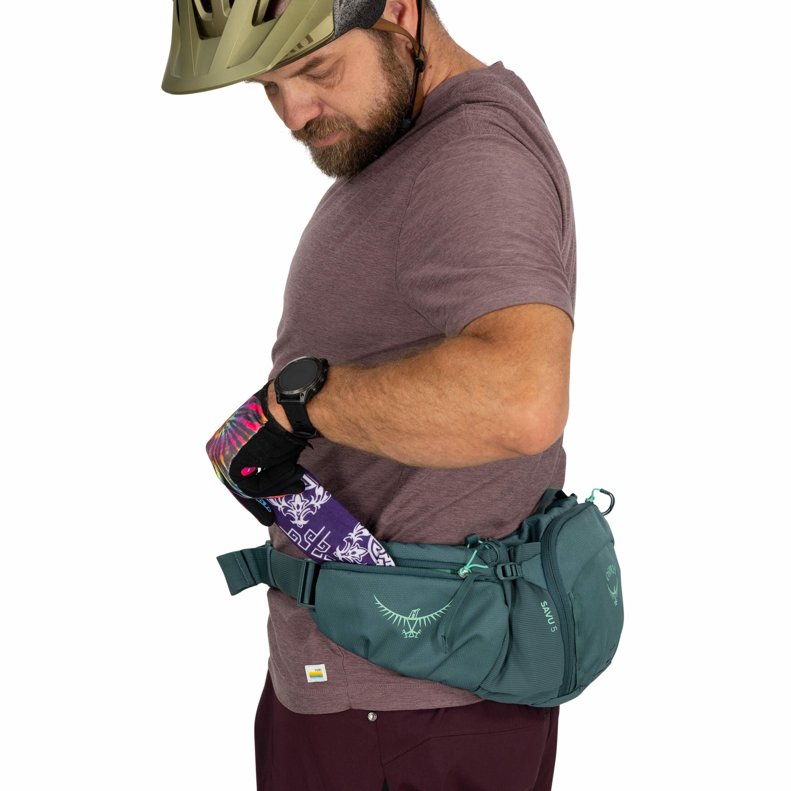 The image shows a person wearing cycling gear, including a helmet and a smartwatch, preparing to place a colorful water bottle into a teal-colored Osprey hip bag. The bag is secured around the person's waist with visible logos of Osprey and the word "SAVU