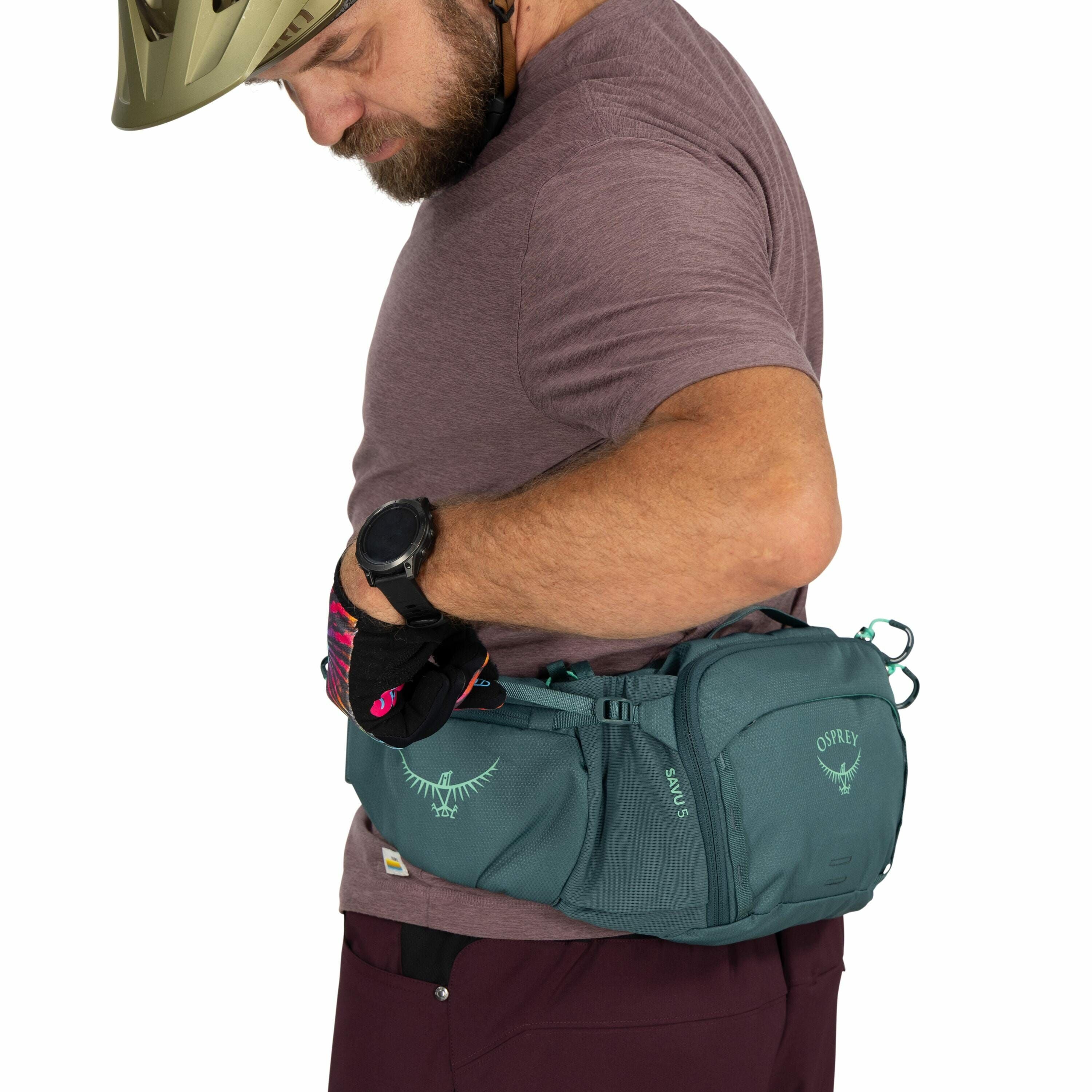 The image shows a man wearing a helmet and dressed casually in a short-sleeve, purple shirt with maroon pants. He has a large Osprey branded fanny pack secured around his waist, prominently featuring its logo and the model name "Savu S." The fanny pack is