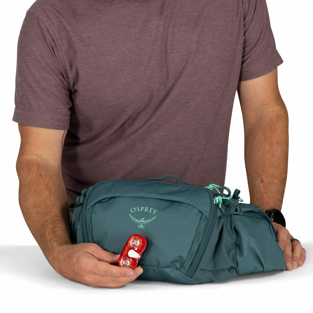The image features a man wearing a short-sleeved, maroon shirt, with his forearms visible. He is holding a teal-colored Osprey branded backpack with both hands. In his left hand, he is holding a small, red object with two lights, which appears to be a bic
