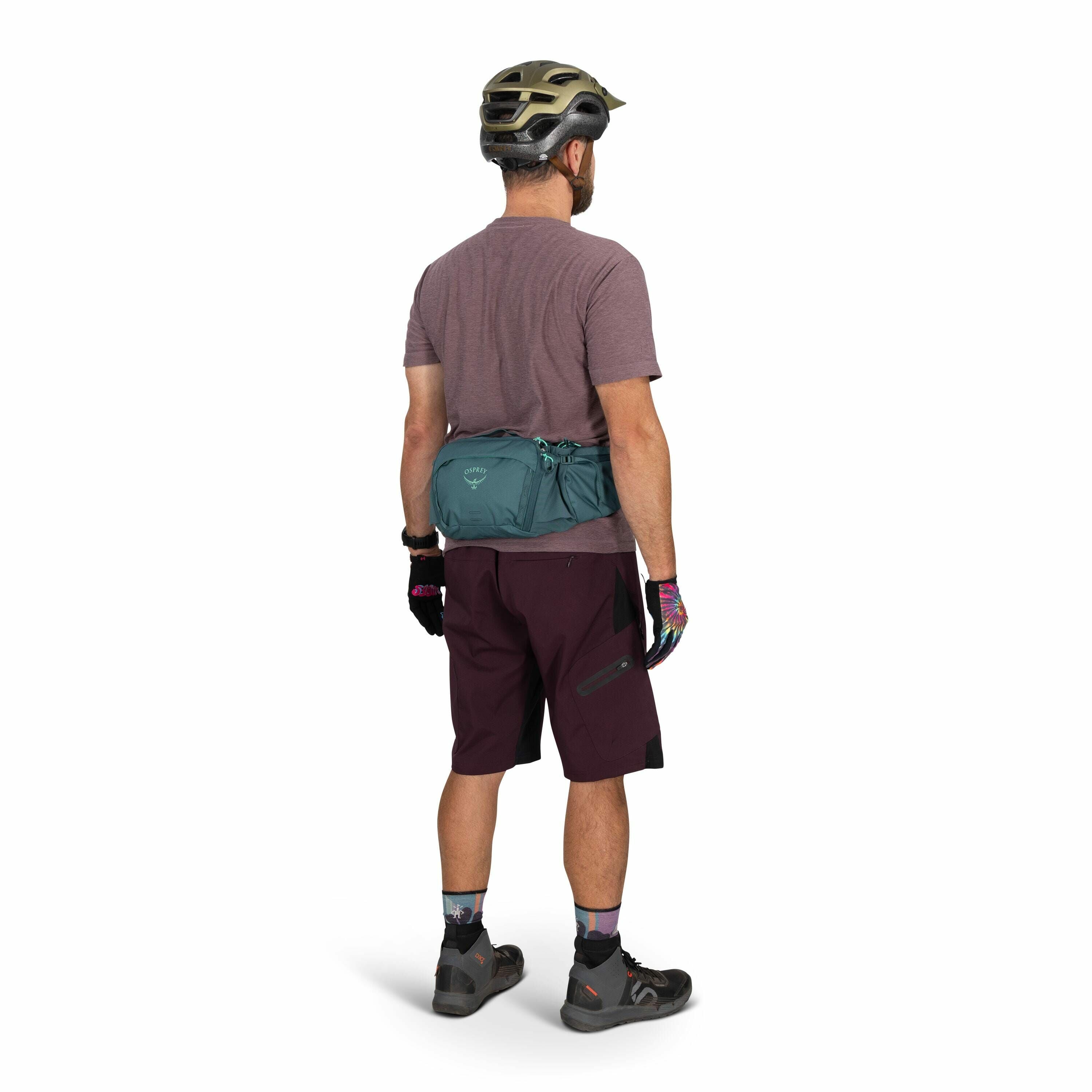 The image shows a man standing with his back to the camera, dressed in mountain biking attire. He is wearing a purple t-shirt and dark purple cycling shorts with black accents, along with colorful gloves and socks. On his head is a protective biking helme