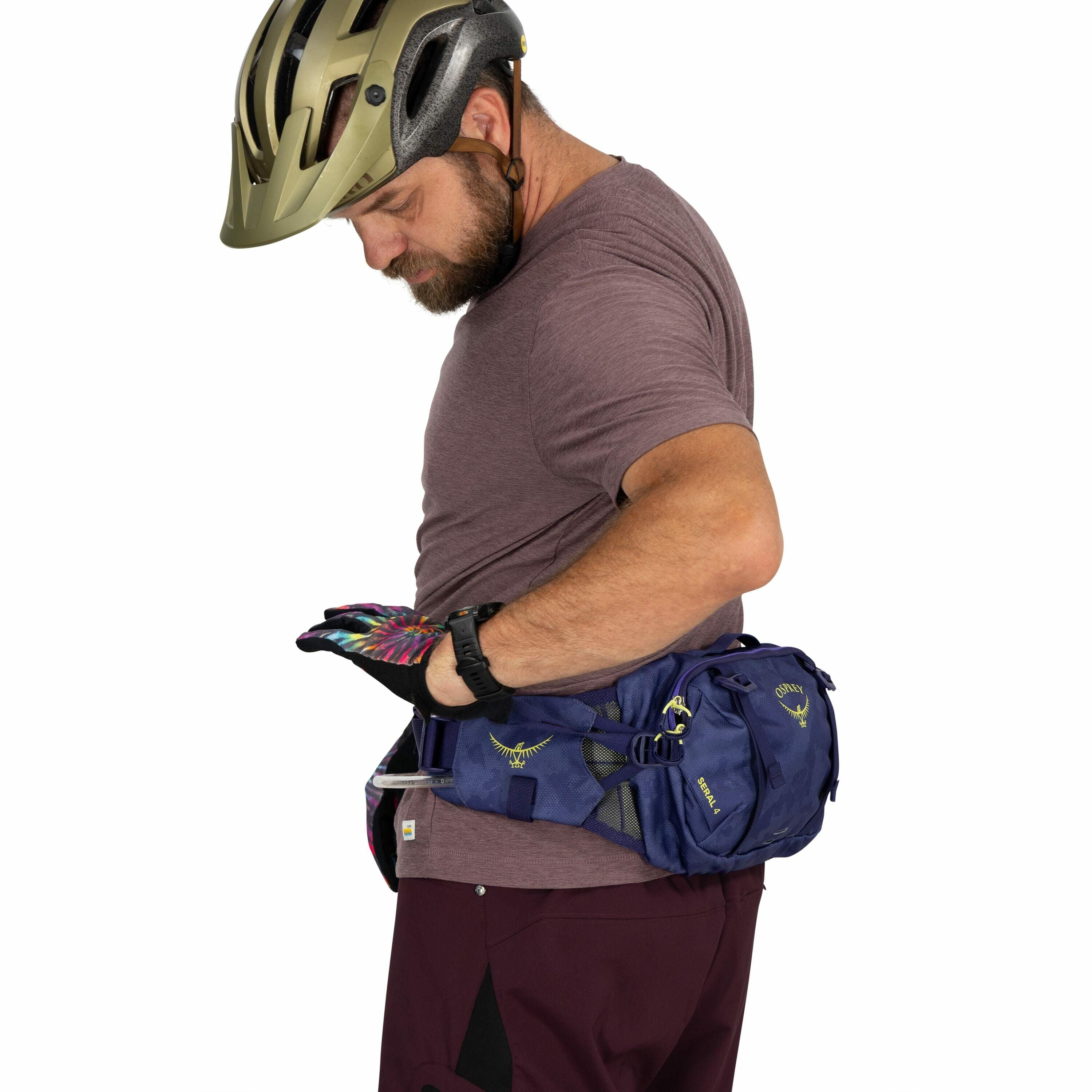 The image shows a man wearing a bicycle helmet and a short-sleeved maroon shirt. He is adjusting or checking the contents of a blue Osprey waist pack secured around his waist. The waist pack has a logo on it and appears to be designed for outdoor activiti