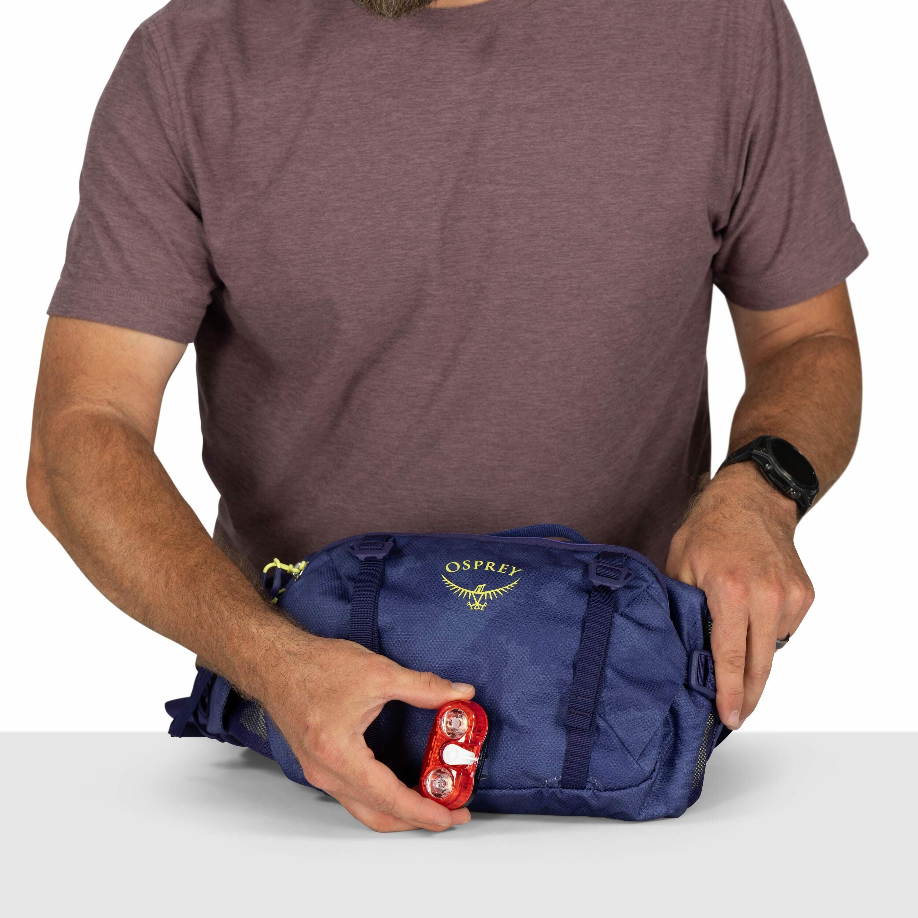 The image shows a man wearing a maroon t-shirt and a black smartwatch on his left wrist. He is holding a blue Osprey bag with one hand while using the other hand to attach a red light device to the bag. The Osprey logo, featuring a stylized bird, is visib