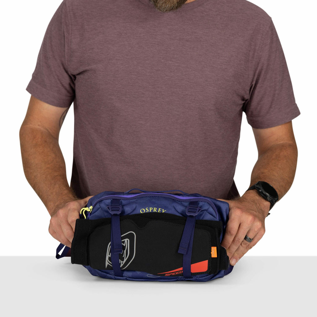 The image features a man with a beard, wearing a short-sleeved maroon t-shirt, standing against a white background. He is holding a blue Osprey bag, which has a visible logo on it. Strapped to the bottom of the bag is what appears to be a black knee or el