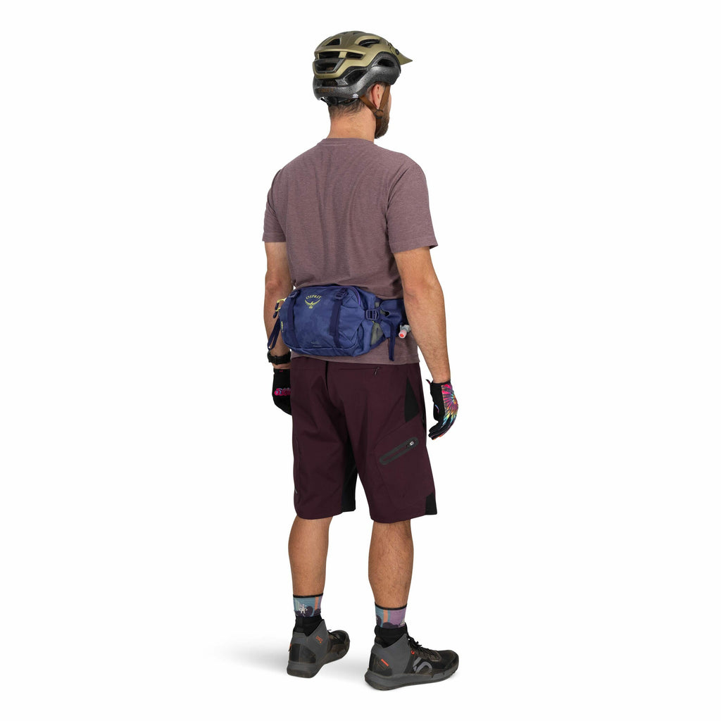 The image shows a man dressed in cycling gear, viewed from the back. He is wearing a dark gray and gold helmet, a purple t-shirt, burgundy cycling shorts, and black cycling shoes with gray accents. On his hands are multicolored gloves, and around his wais