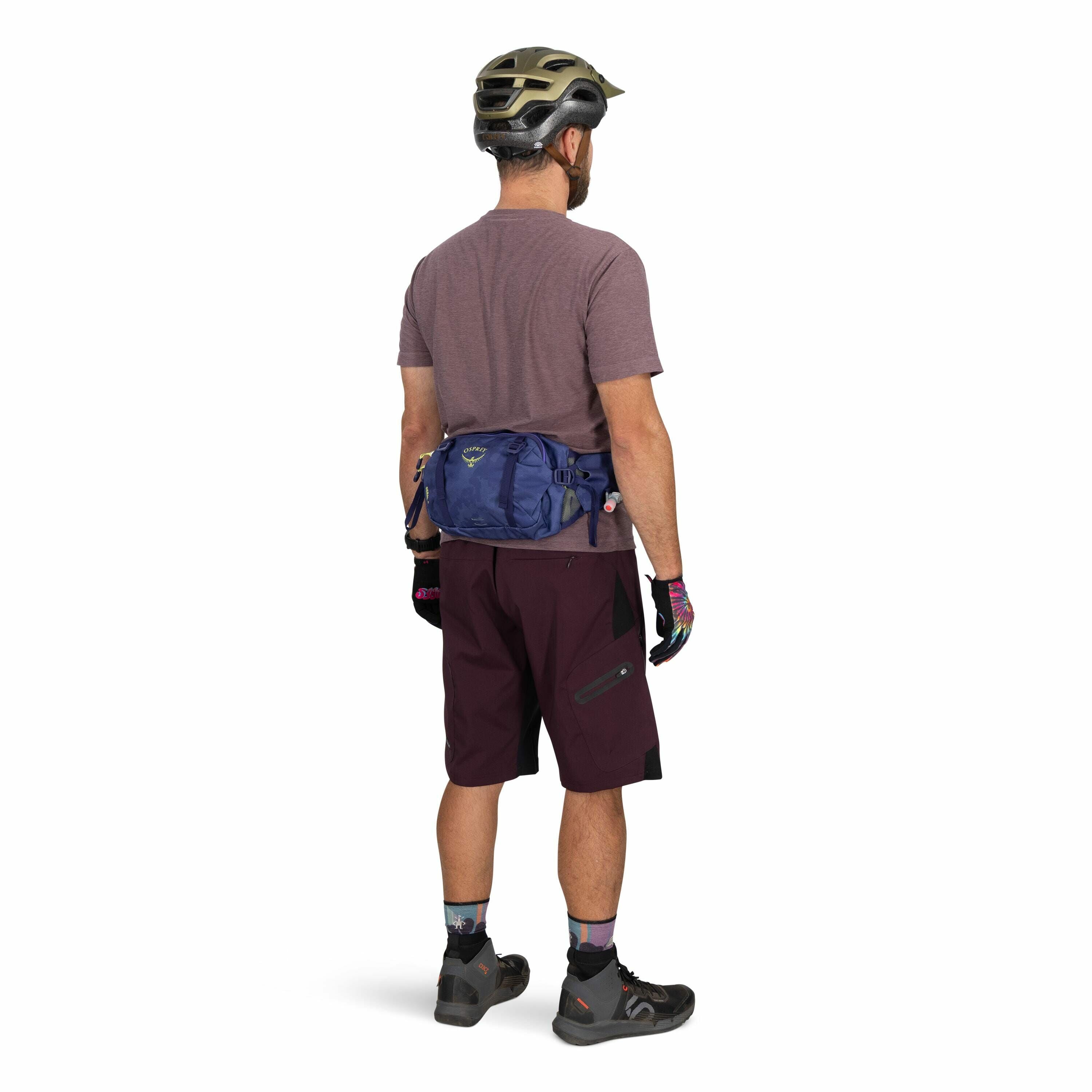 The image shows a man dressed in cycling gear, viewed from the back. He is wearing a dark gray and gold helmet, a purple t-shirt, burgundy cycling shorts, and black cycling shoes with gray accents. On his hands are multicolored gloves, and around his wais
