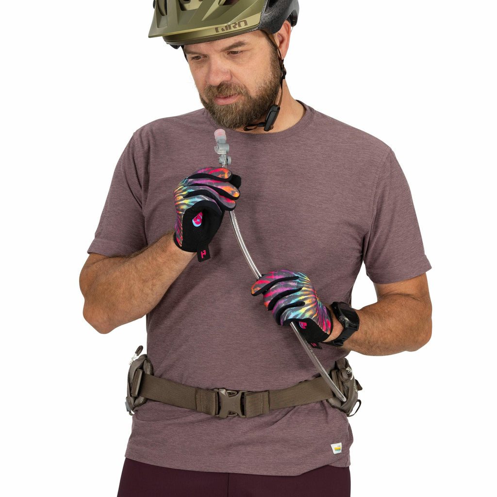 The image shows a person wearing a light purple t-shirt, a green helmet, and colorful gloves that feature a tie-dye pattern with shades of purple, blue, and orange. The person is holding a hydration tube, presumably from a hydration pack, with both hands;