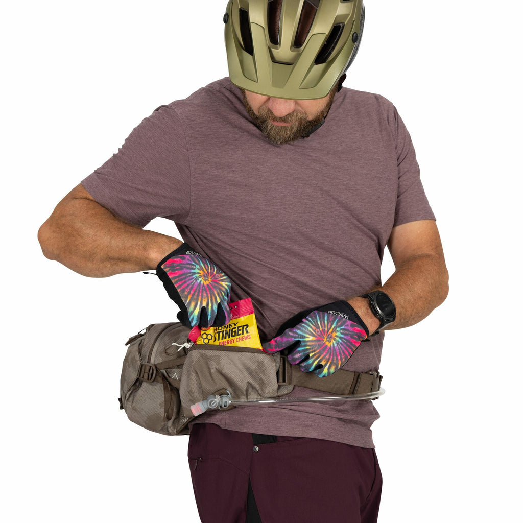 The image depicts a person wearing a helmet and a short-sleeved maroon shirt, looking down as they retrieve an item from a gray waist pack. The person is wearing colorful gloves with a tie-dye pattern, and a smartwatch on their left wrist. They are pullin