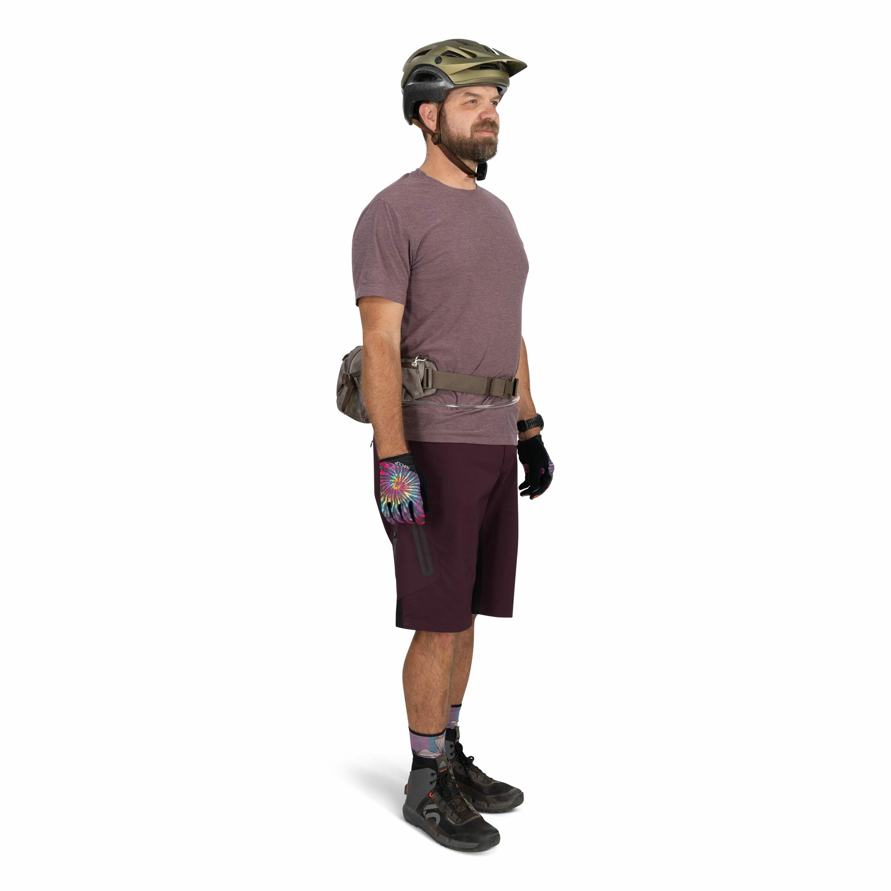 The image depicts a person dressed in casual biking attire standing in a side profile pose. They are wearing a green helmet, a maroon T-shirt, and matching maroon shorts. The individual is equipped with colorful gloves displaying a vibrant tie-dye pattern