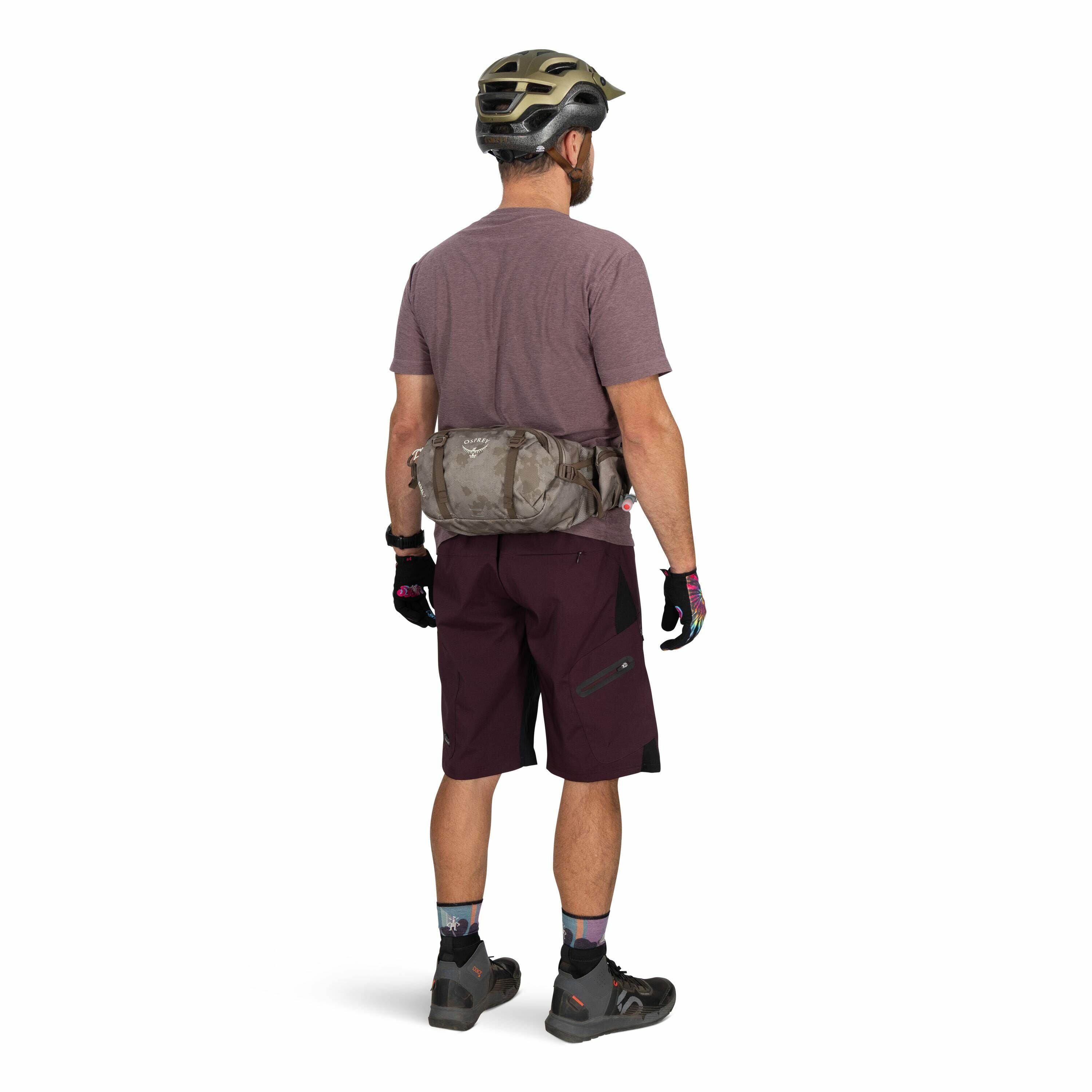 The image shows a person standing with their back facing the camera, dressed for a cycling activity. The individual is wearing a maroon t-shirt and dark maroon shorts that feature pockets with zippers and a panel design. On their back is a camouflaged gra