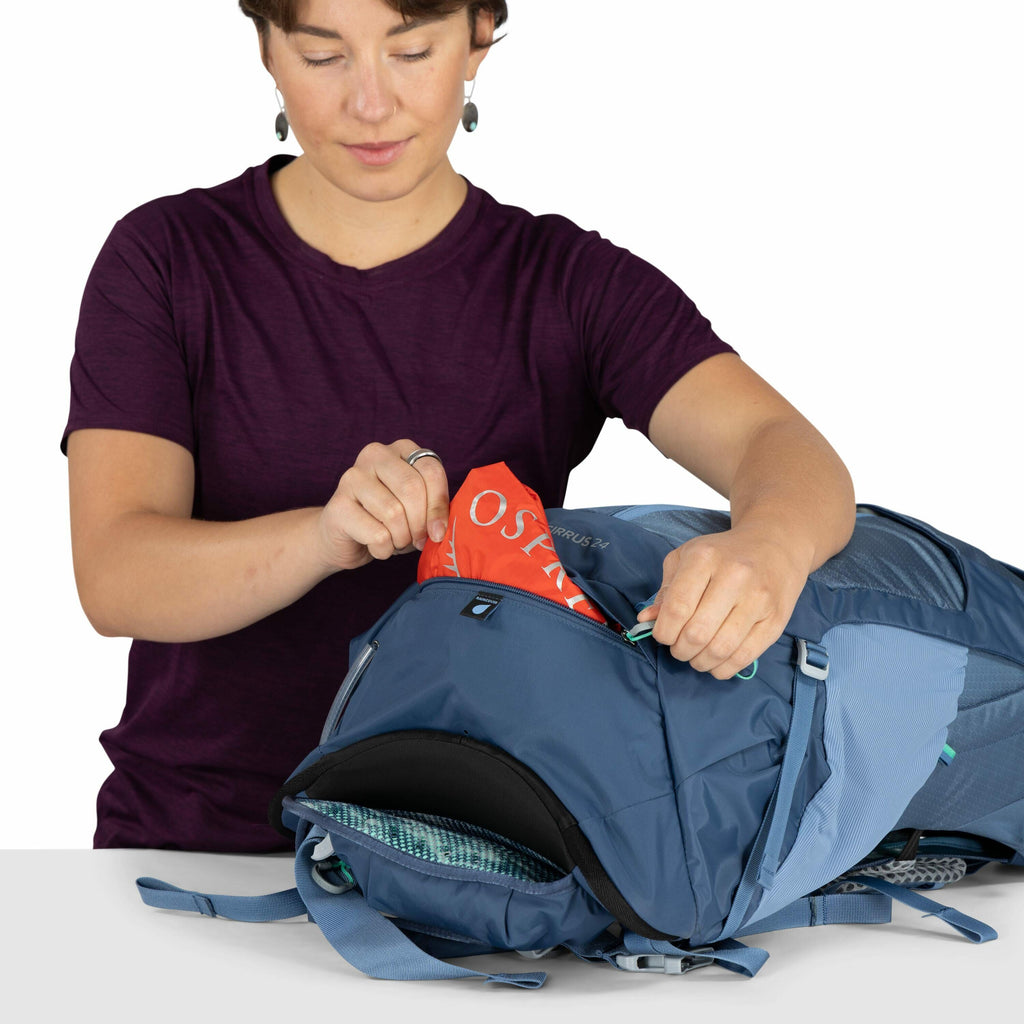 The image shows a person dressed in a dark purple shirt standing behind a table while interacting with a blue backpack. They are holding the zipper open and removing an item that appears to be a rain cover with the word "OSPREY" visible on it, suggesting