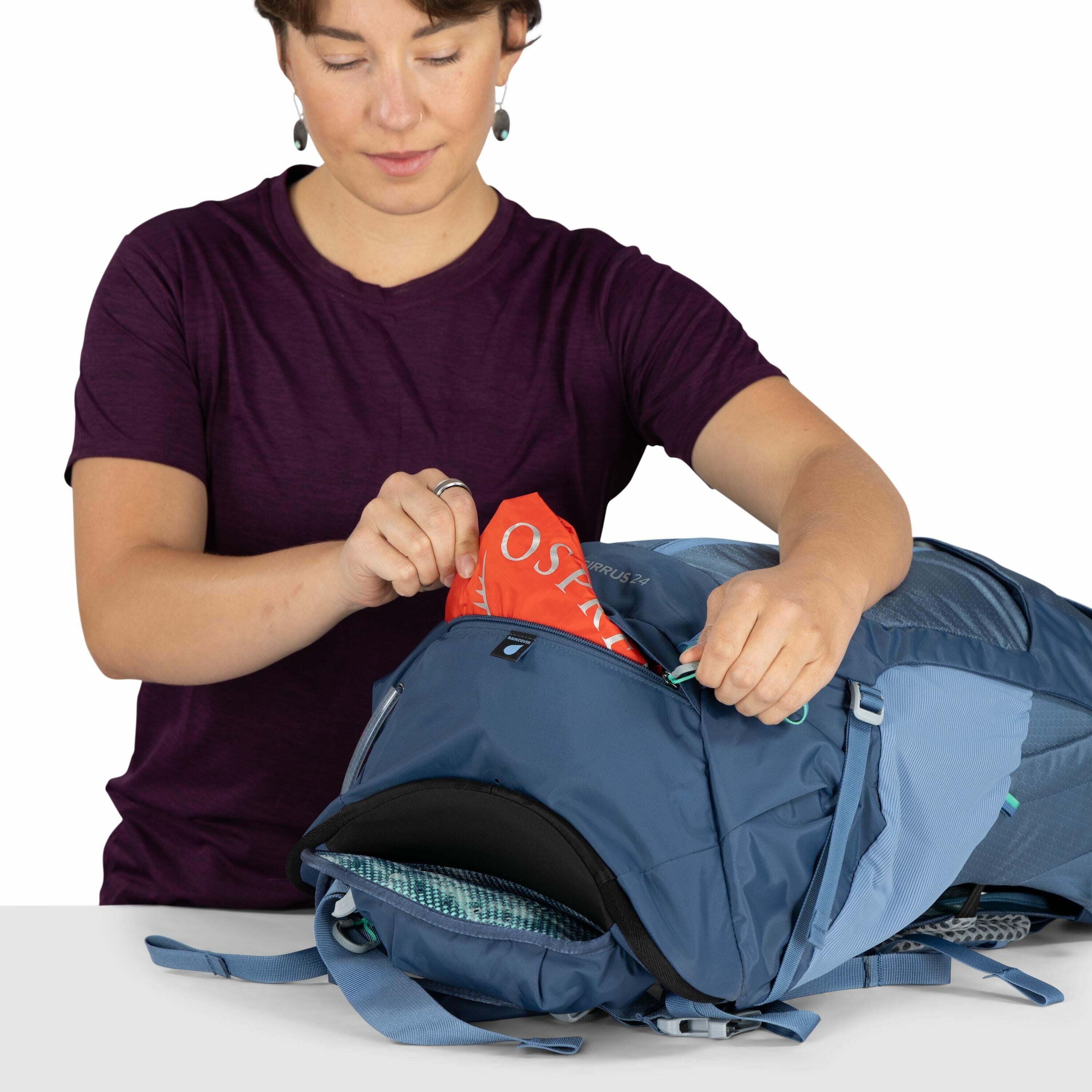 The image shows a person dressed in a dark purple shirt standing behind a table while interacting with a blue backpack. They are holding the zipper open and removing an item that appears to be a rain cover with the word "OSPREY" visible on it, suggesting