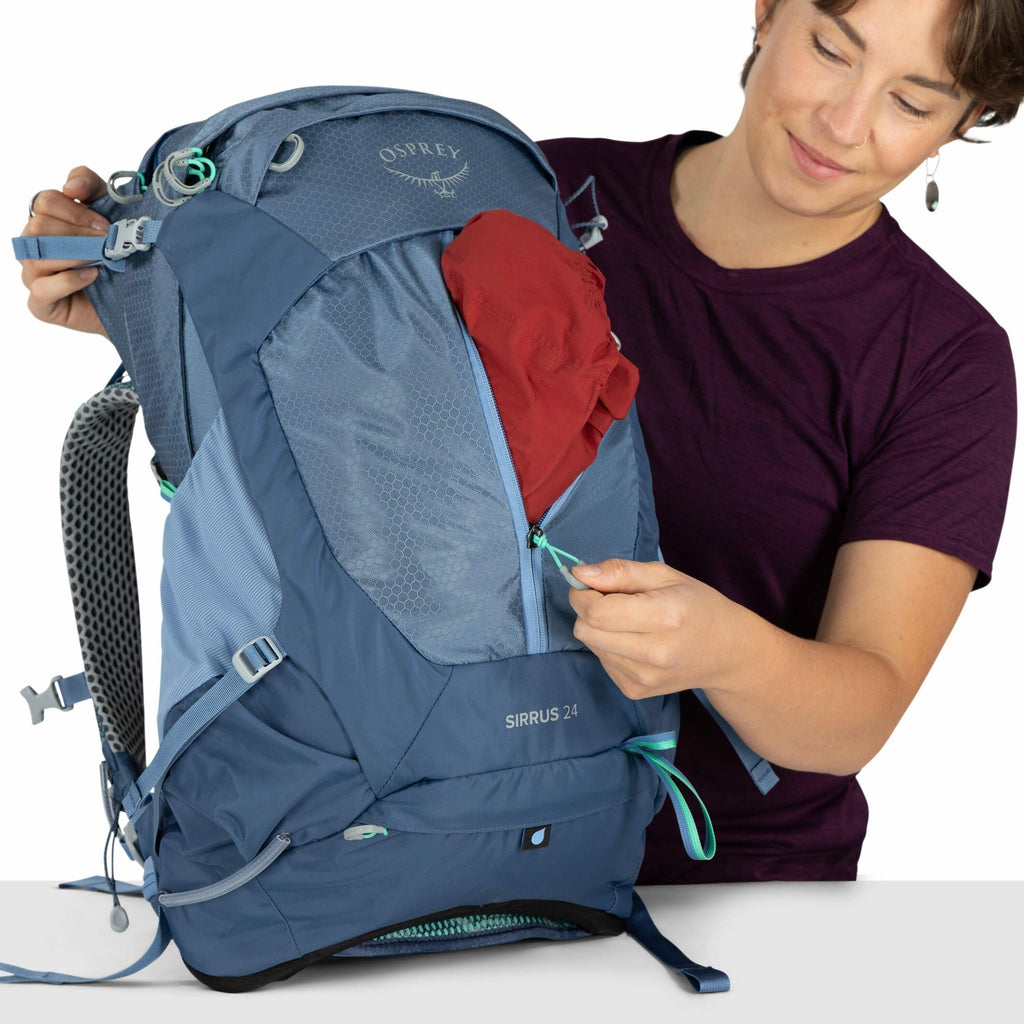 The image shows a person dressed in a dark purple t-shirt holding a blue Osprey backpack model "Sirrus 24". The individual appears to be opening a front compartment of the backpack, which contains a red item, possibly clothing. The backpack has various fe