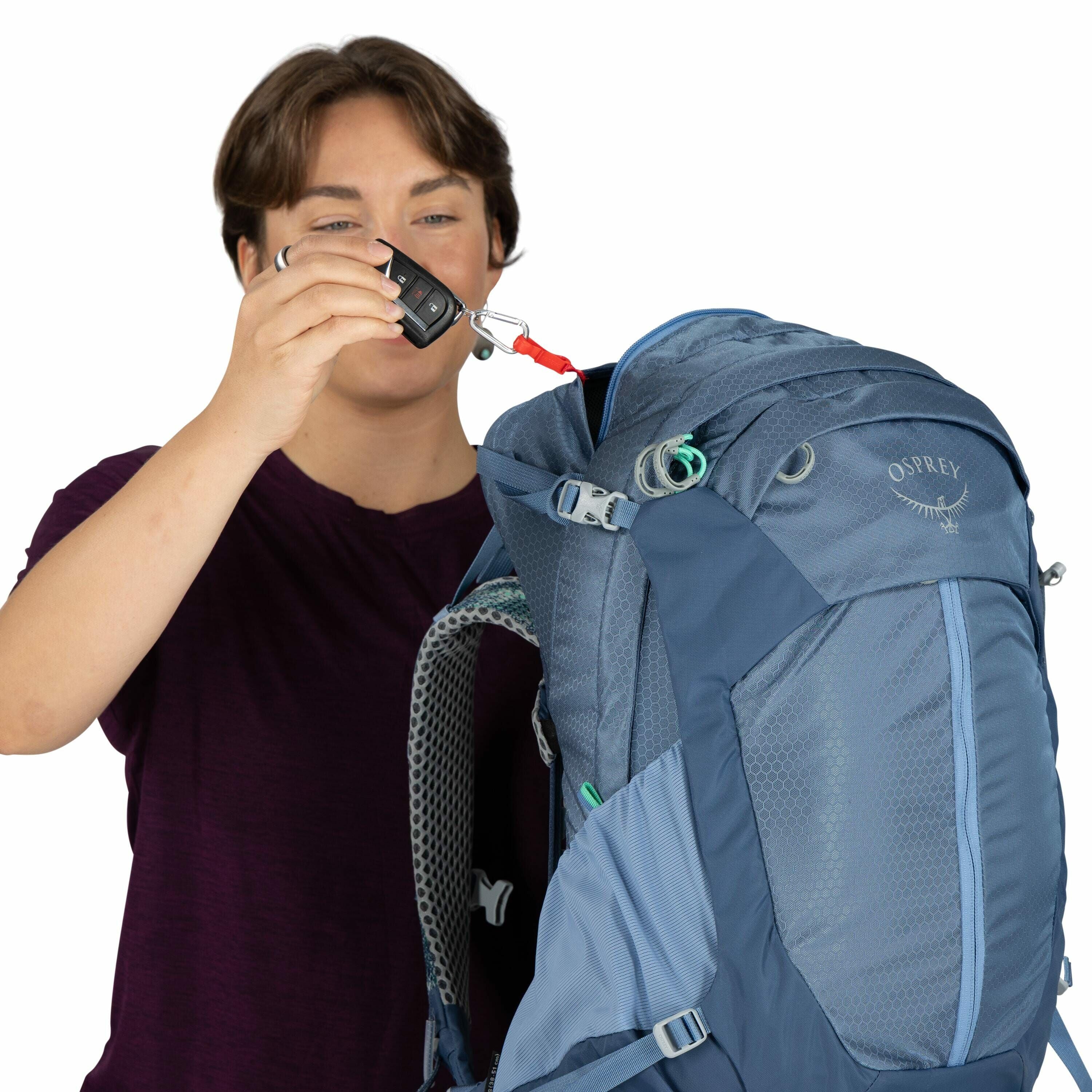 The image shows a person holding an Osprey backpack. The backpack is primarily blue with a combination of darker and lighter blue shades, featuring a patterned texture on some sections. It has several adjustable straps and buckles, along with a visible lo