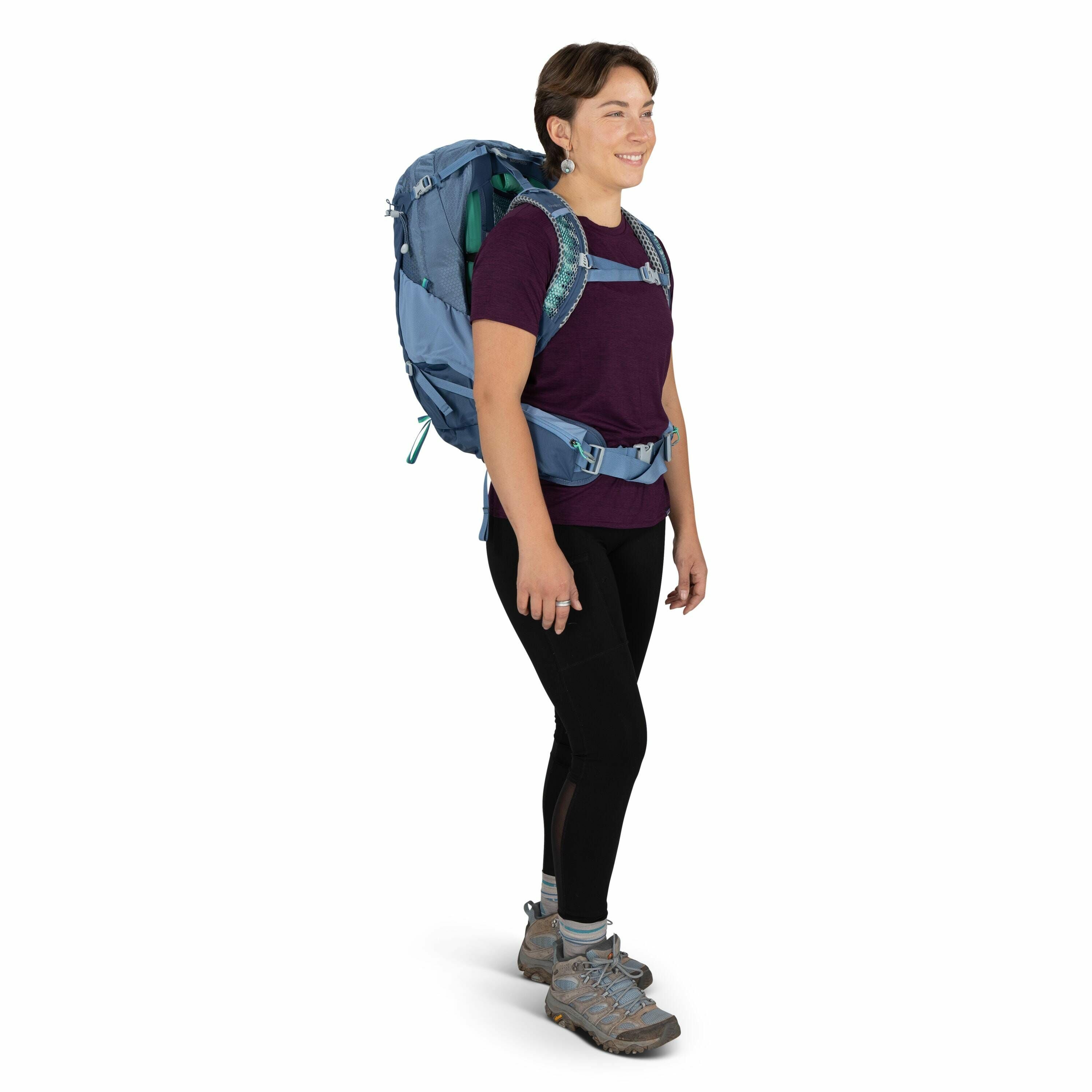 The image depicts a person standing and wearing outdoor hiking gear. They have a large blue backpack with multiple compartments and straps, suggesting it is equipped for long hikes or treks. The person is dressed in a short-sleeved, purple top paired with