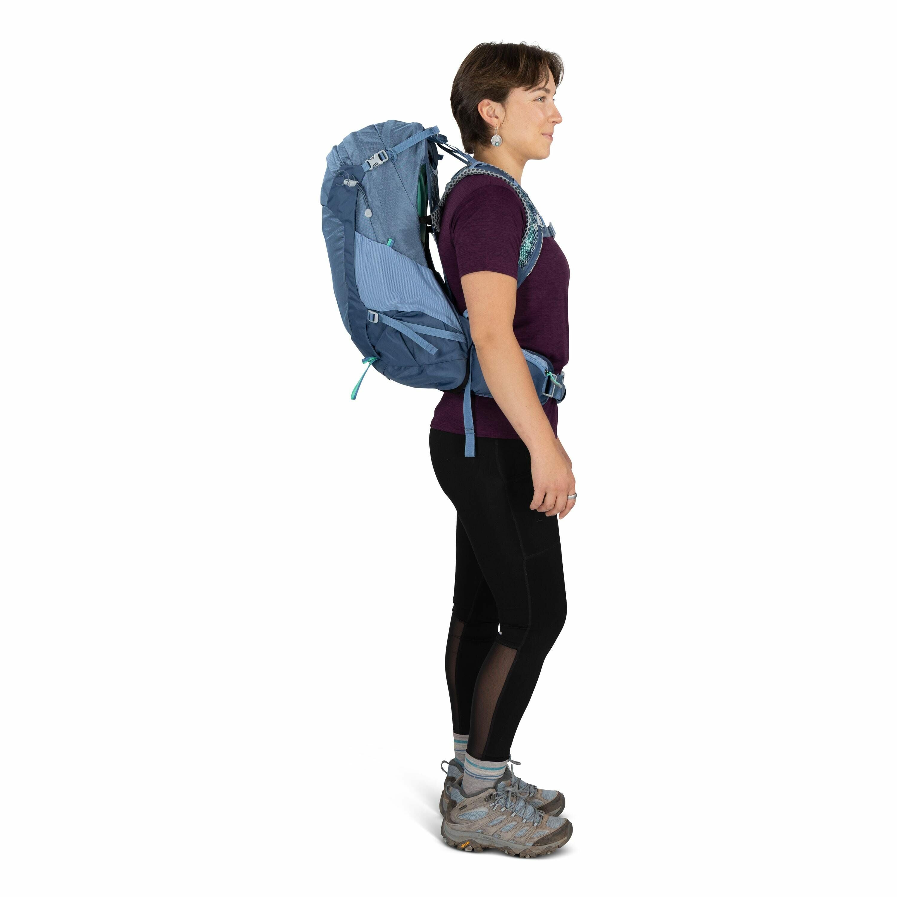 The image shows a person standing sideways, wearing outdoor gear. They have a large blue hiking backpack with various straps secured across the shoulders and waist, suggesting readiness for outdoor activities or travel. The person is dressed in a dark pur