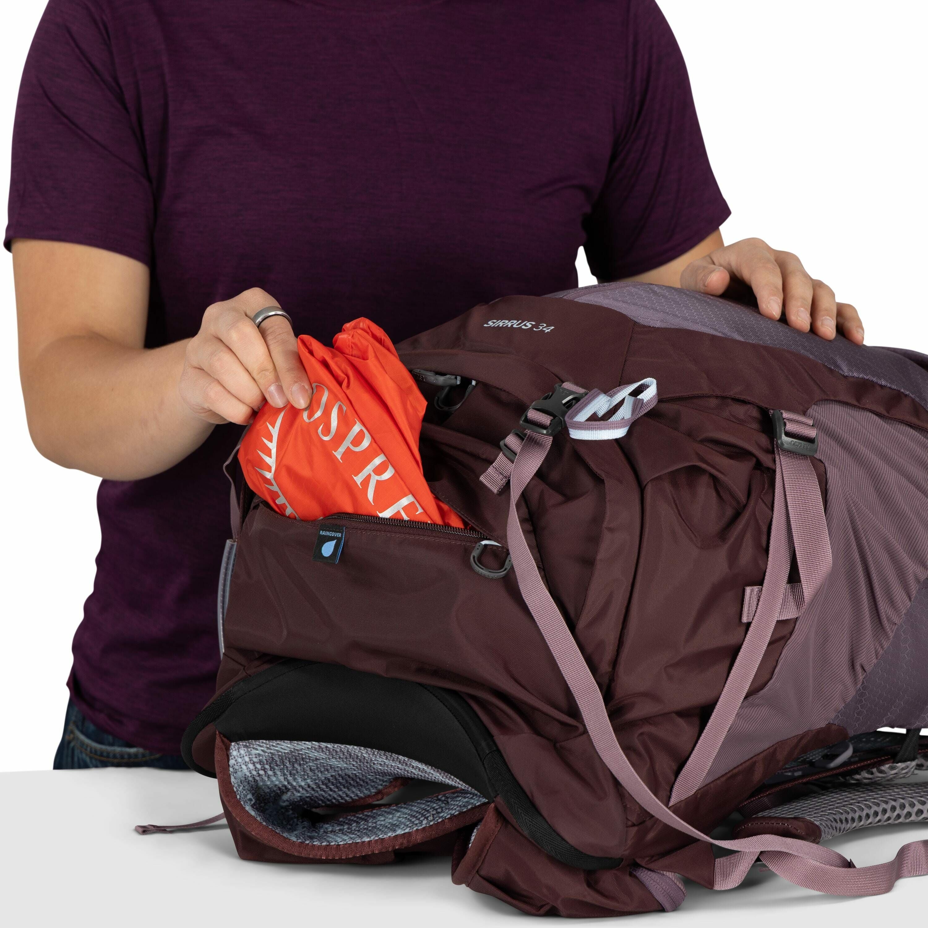 The image shows a person wearing a short-sleeved, dark purple shirt, holding a backpack with one hand while placing or retrieving an item with the other. The backpack is primarily maroon, with some light gray details and straps, and the brand name "SIRRUS