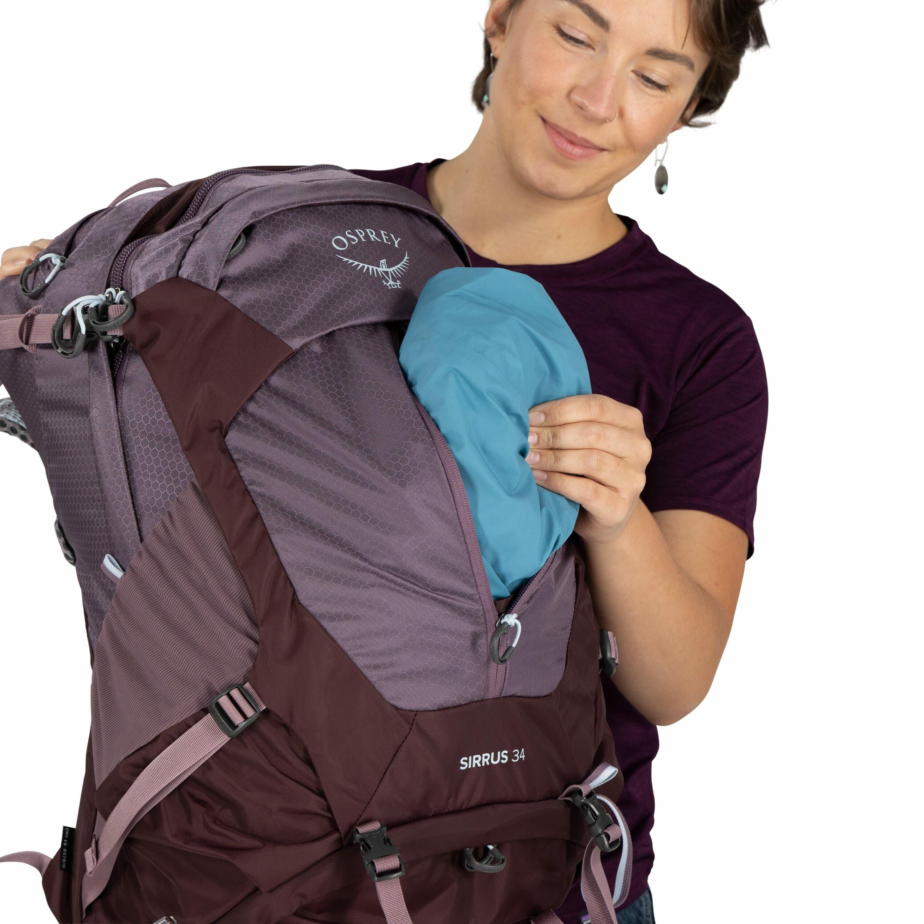 The image shows a person holding a hiking backpack made by Osprey. The backpack is a two-tone color, combining shades of purple and brown, and has the model name "SIRRUS 34" printed on it. The person is pulling out or putting away a bright blue item, poss