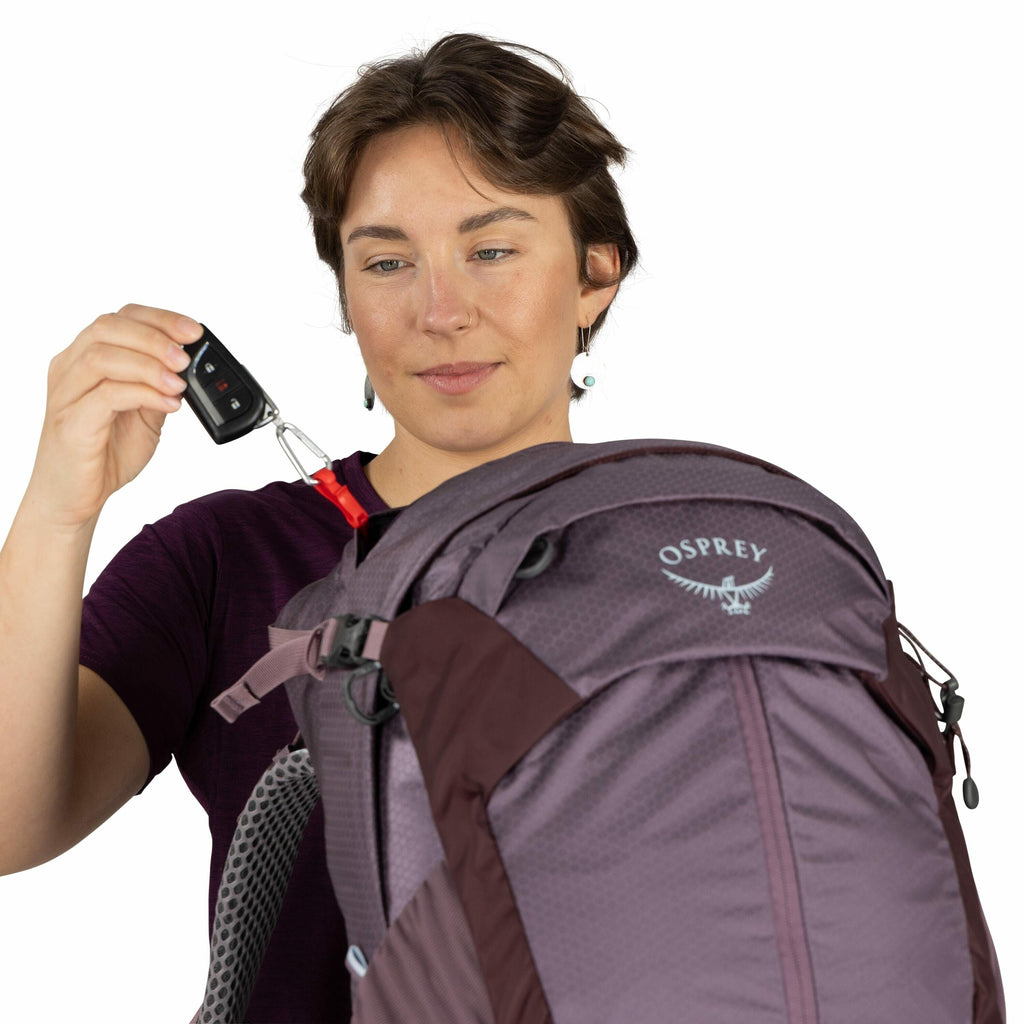 The image shows a person holding a car key fob that is attached to a red carabiner. The carabiner is clipped to a purple Osprey backpack, prominently displaying the brand's logo. The individual is wearing a dark maroon shirt, and their hand is positioned