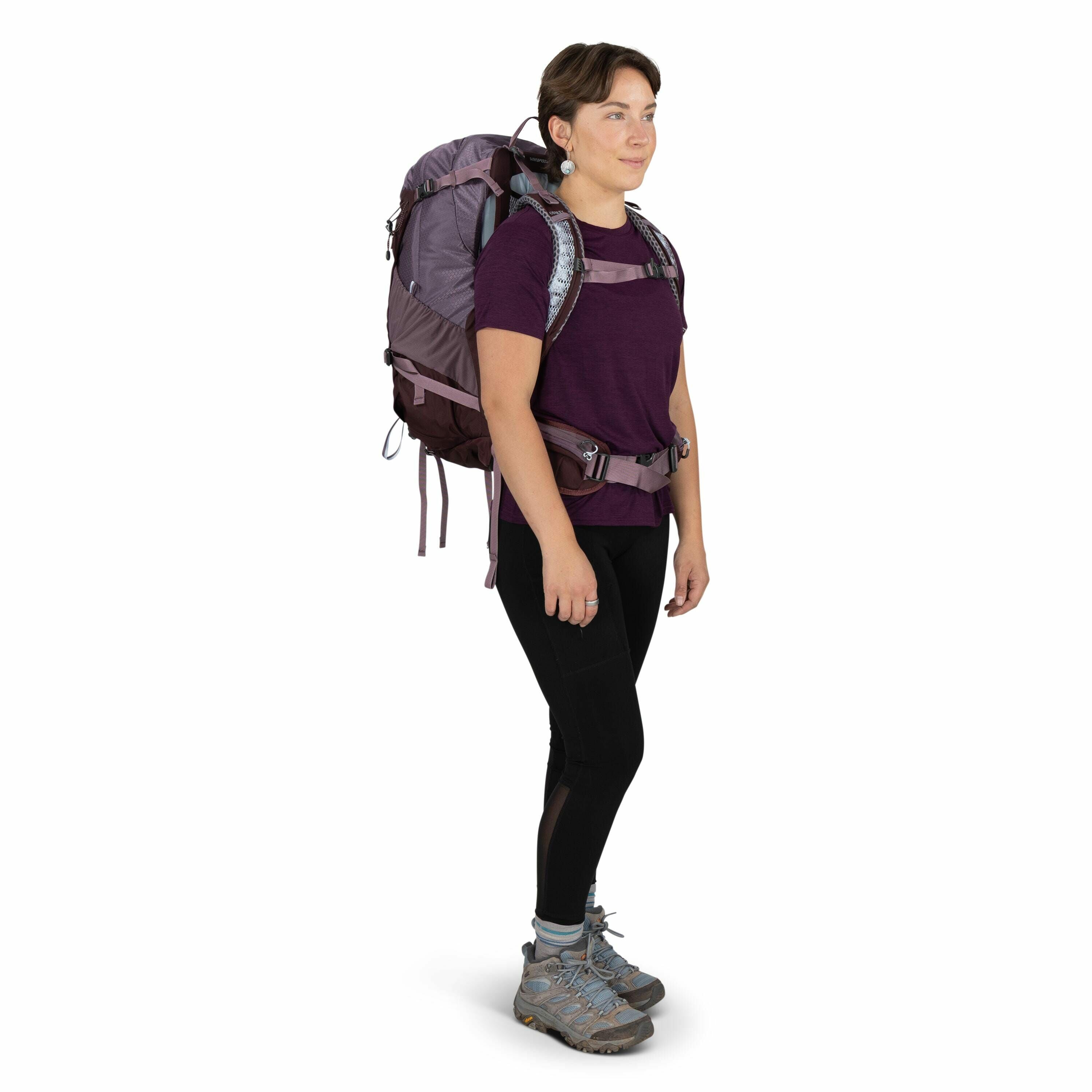 The image shows a person wearing outdoor hiking attire and gear. They are dressed in a purple short-sleeve shirt and black leggings, equipped with a large hiking backpack in shades of purple and gray. The backpack appears well-fitted, with its straps secu