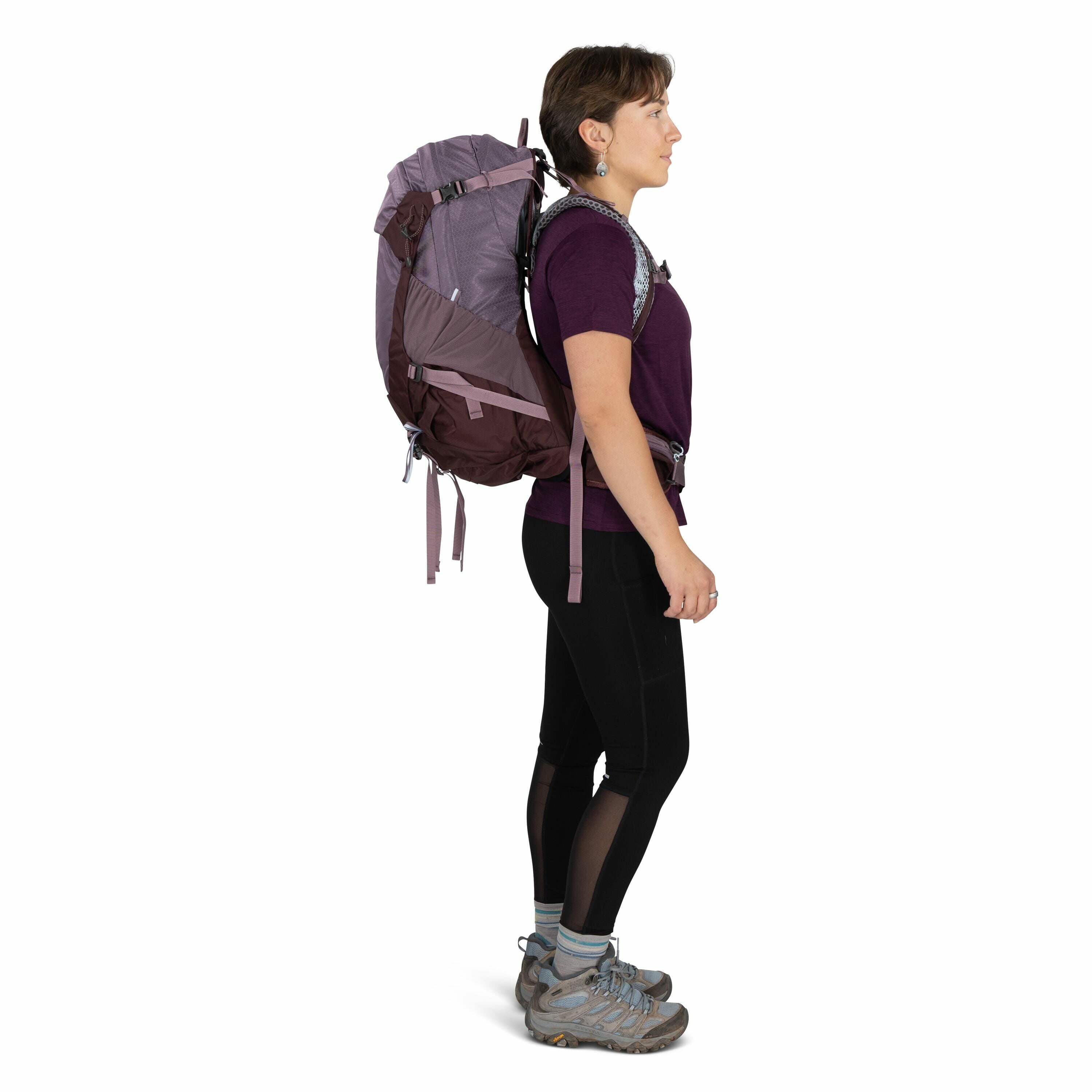 The image shows a person standing in a profile view, wearing outdoor hiking gear. They are dressed in a short-sleeve purple shirt, black leggings with mesh panels, and gray hiking shoes. The individual is carrying a large purple and brown backpack that ha