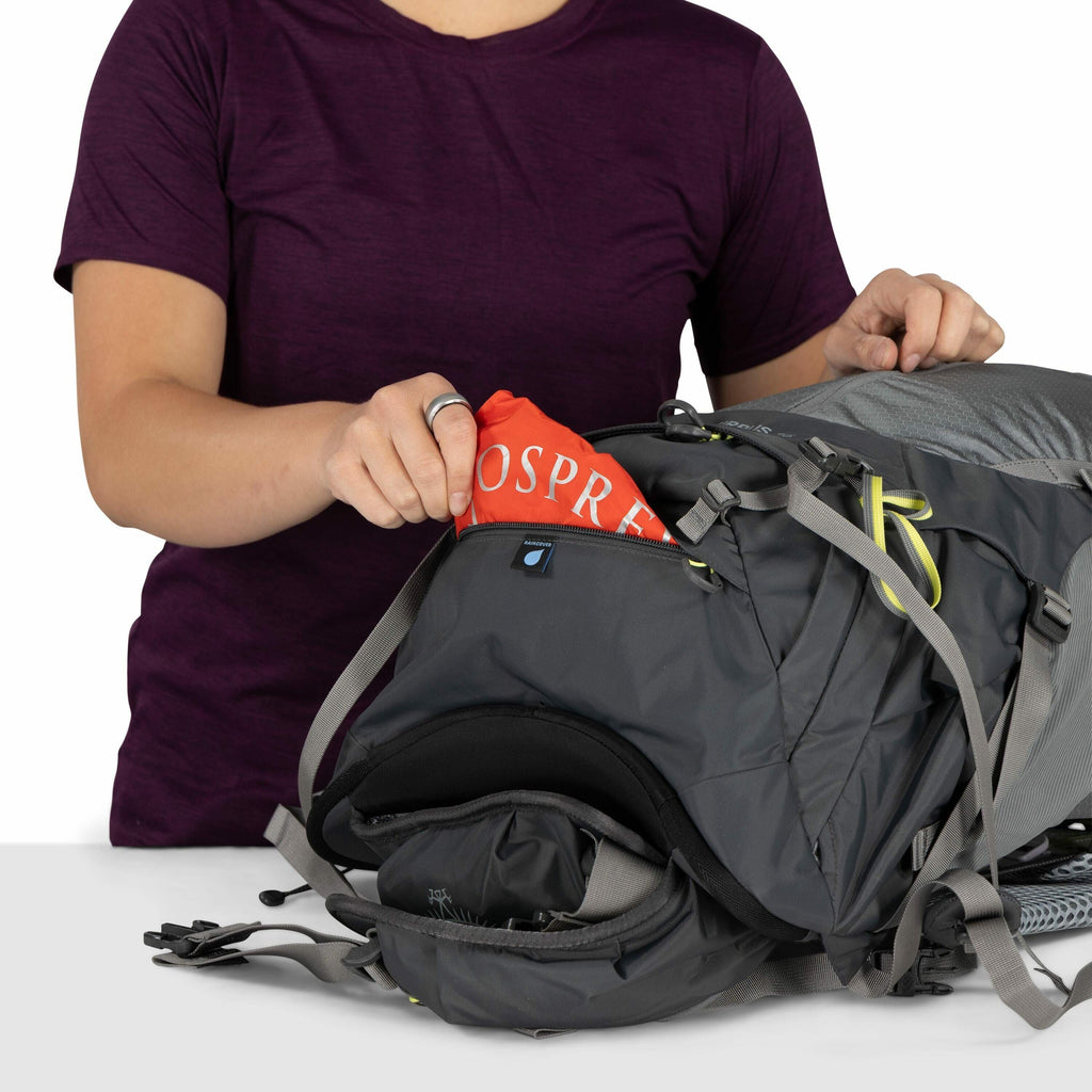 The image shows a person wearing a dark purple shirt, placing an Osprey rain cover into the front pocket of a dark gray backpack. The rain cover is red and prominently displays the Osprey brand name. The backpack appears to be designed for hiking or outdo