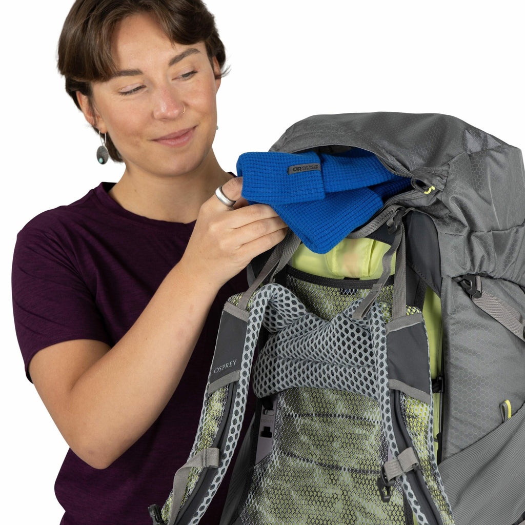 The image shows a person wearing a dark purple shirt, opening a grey backpack. The backpack is an Osprey brand, as indicated by the logo on the shoulder strap. The interior of the backpack features green mesh padding for comfort and ventilation. Inside th