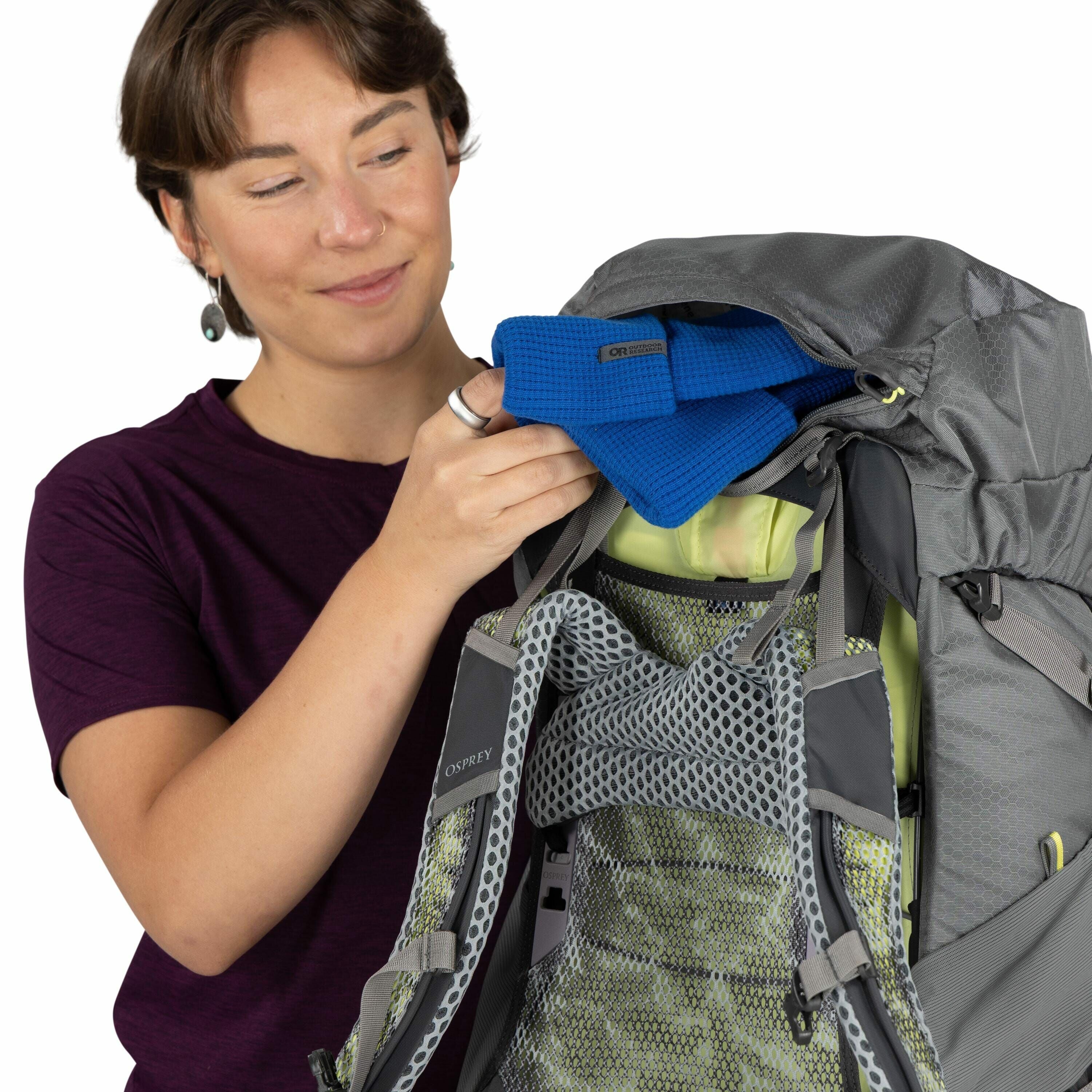 The image shows a person wearing a dark purple shirt, opening a grey backpack. The backpack is an Osprey brand, as indicated by the logo on the shoulder strap. The interior of the backpack features green mesh padding for comfort and ventilation. Inside th