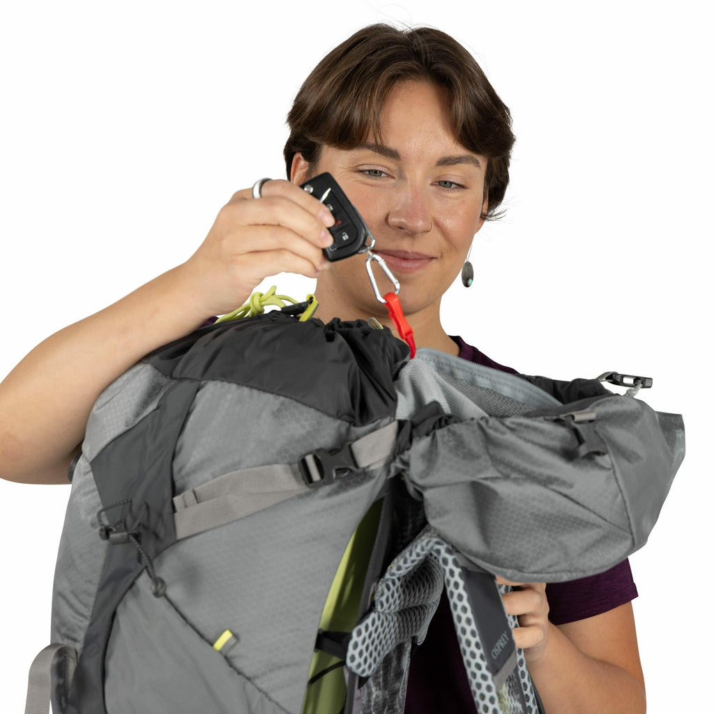 The image depicts a person holding a gray hiking backpack with various compartments and features. The backpack is open at the top, revealing its interior, which includes a red strap and a green internal lining or gear. The person is wearing a purple shirt