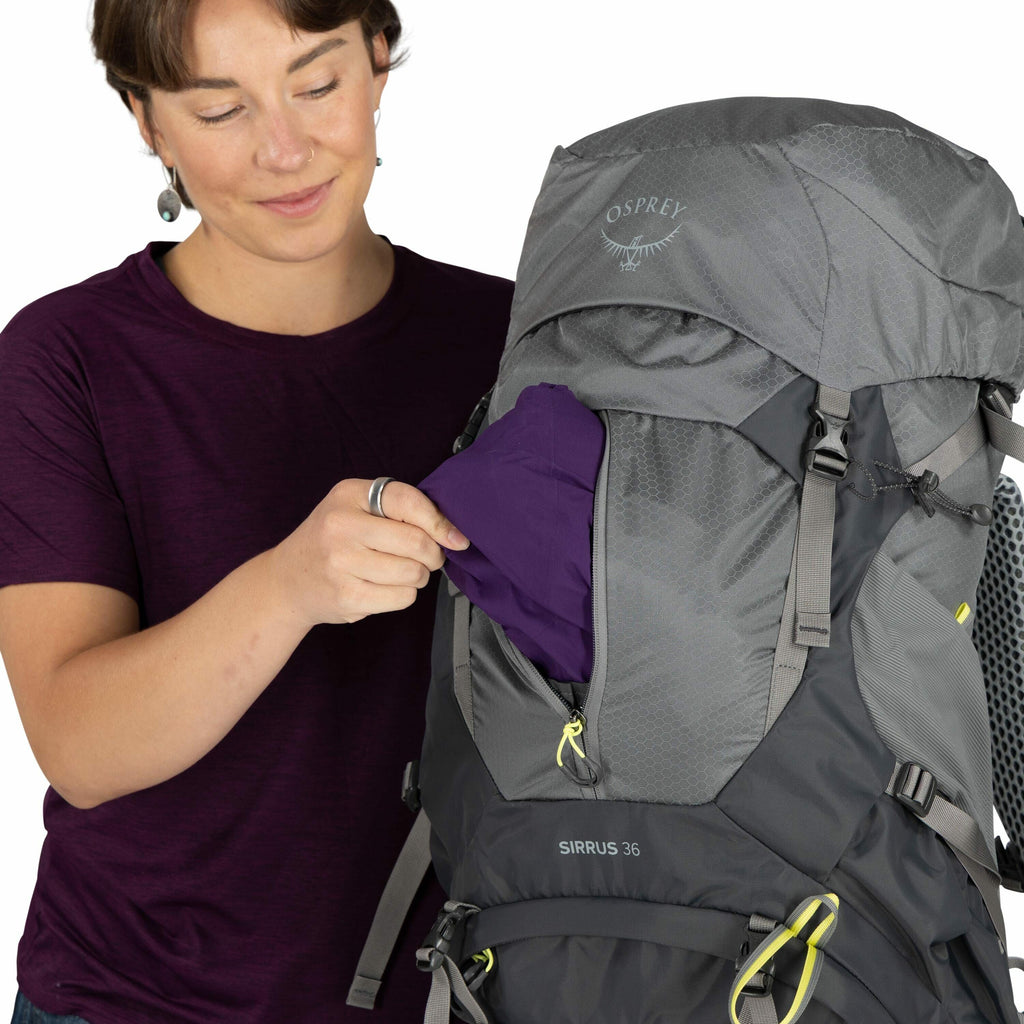 The image shows a person holding a gray and dark-colored Osprey Sirrus 36 backpack. They are in the process of placing or retrieving a purple fabric item from the top compartment of the backpack, which is partially unzipped. The person is wearing a dark p