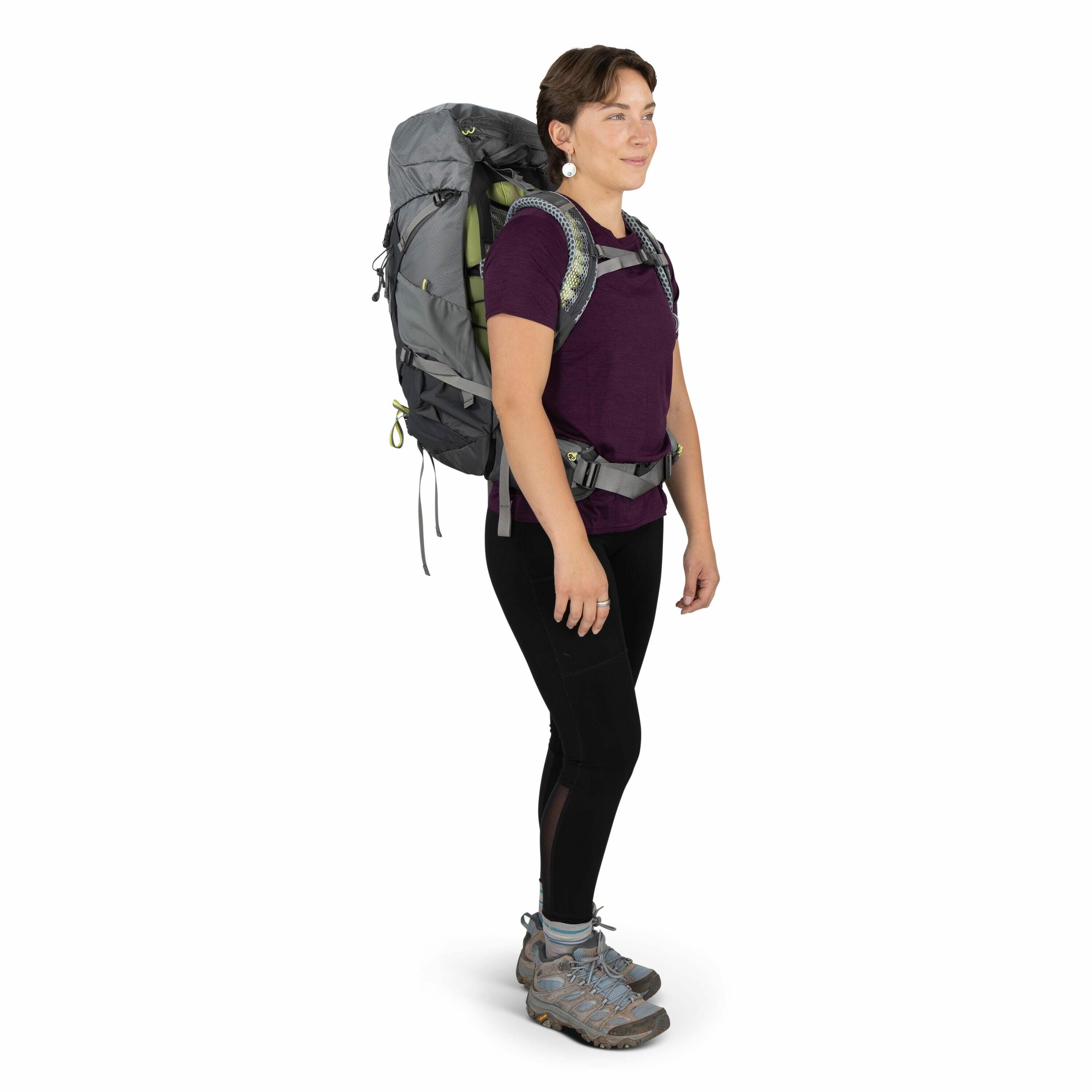 The image shows a person standing in profile, wearing hiking gear. They are dressed in a purple t-shirt and black leggings, and they have on sturdy grey hiking boots with blue accents. The individual is carrying a large grey and green backpack, which is e