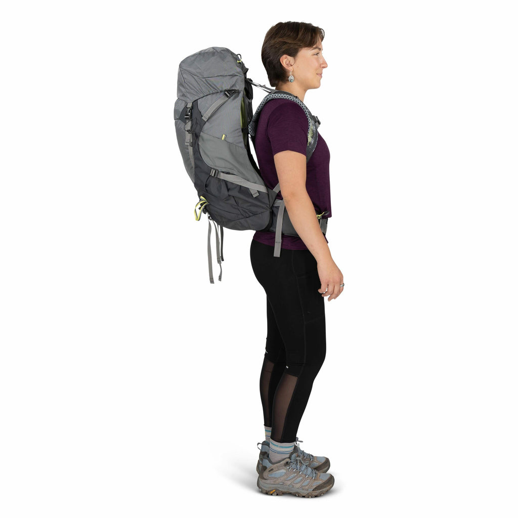 The image depicts a person standing in profile view, equipped for hiking or trekking. They are wearing a large gray backpack that appears to be well-fitted with multiple straps for support. The individual is dressed in a purple short-sleeve shirt and blac