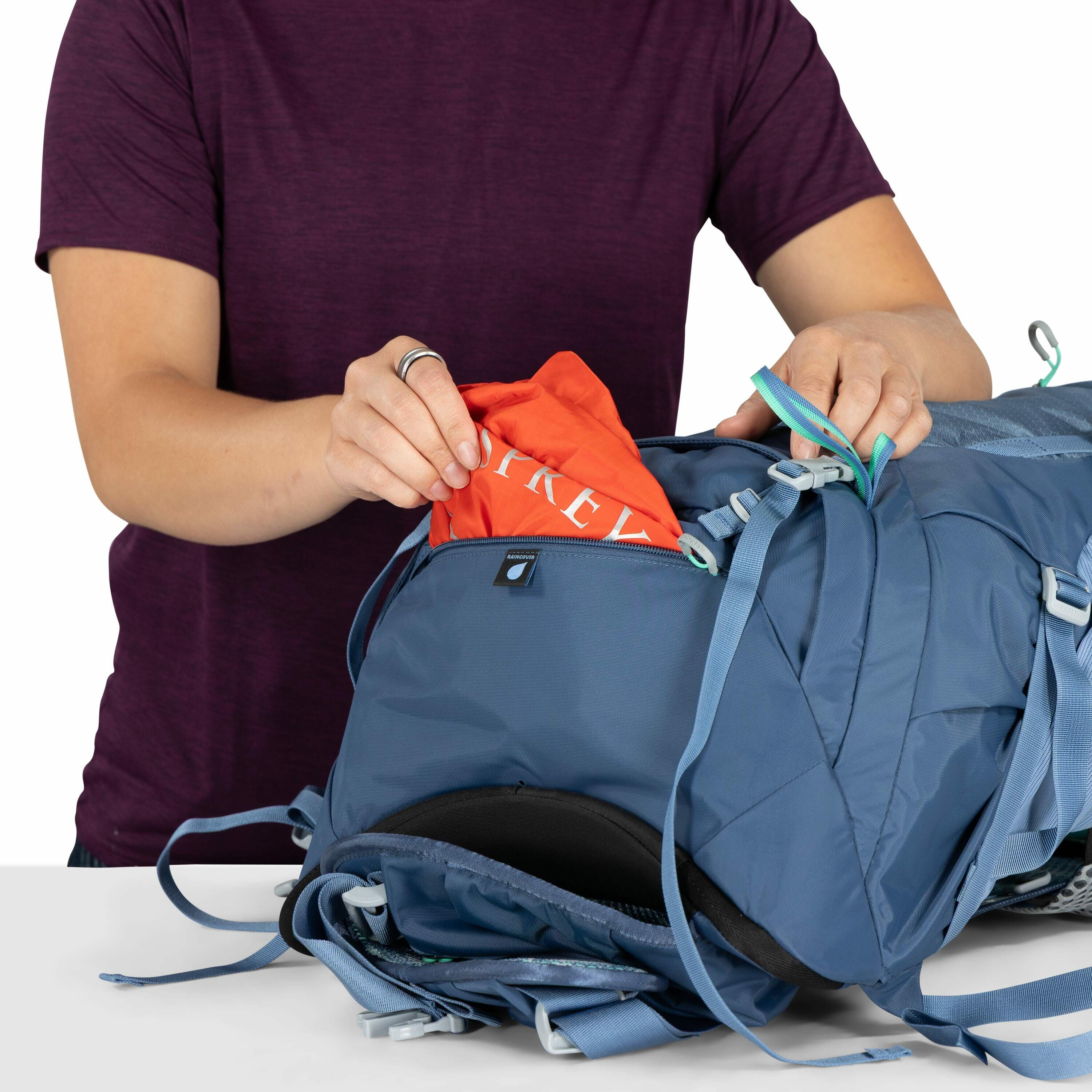 The image shows a person wearing a dark purple t-shirt as they are in the process of packing or unpacking a blue backpack. The individual's hands are placing an item of clothing or fabric, which is orange and has the word "OSPREY" printed on it, into the