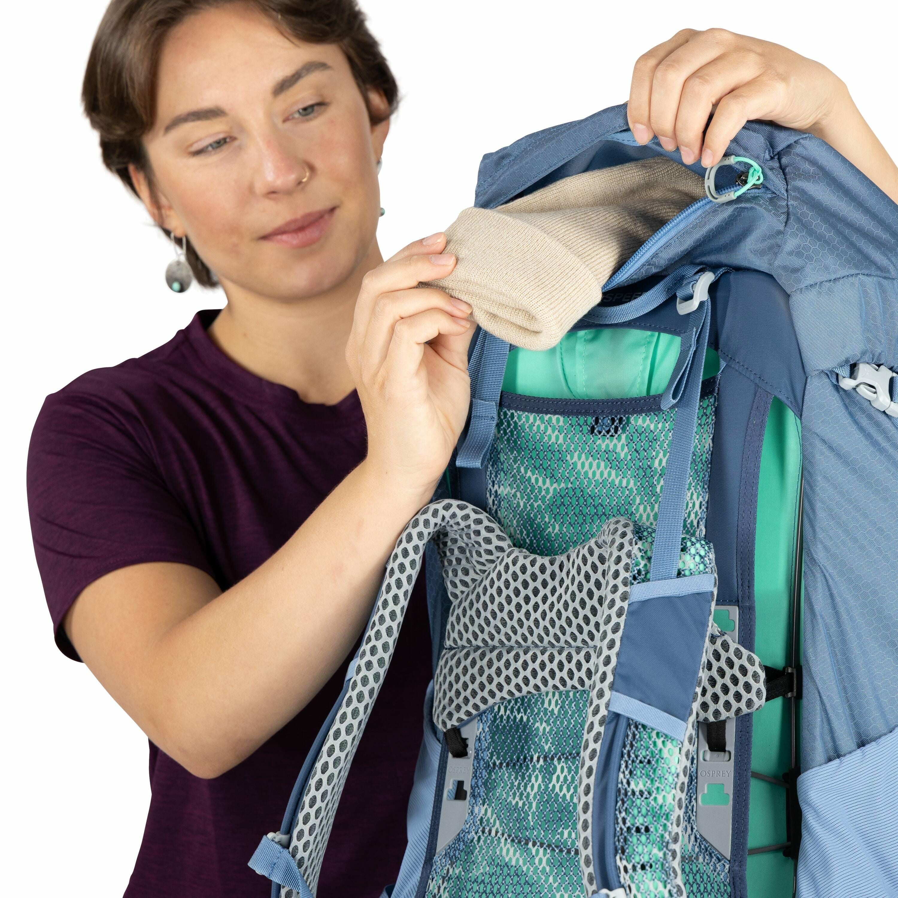 The image shows a person demonstrating a blue backpack with gray and turquoise accents. The person is holding the top of the backpack open and inserting a rolled-up beige item, possibly a shirt or towel, into the compartment. The backpack features a mesh
