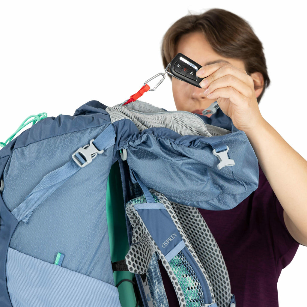 The image shows a person holding a blue Osprey backpack. The backpack appears to be large, suitable for hiking or backpacking, with adjustable straps and mesh padding for comfort and ventilation. The person is lifting the top flap of the backpack with a r