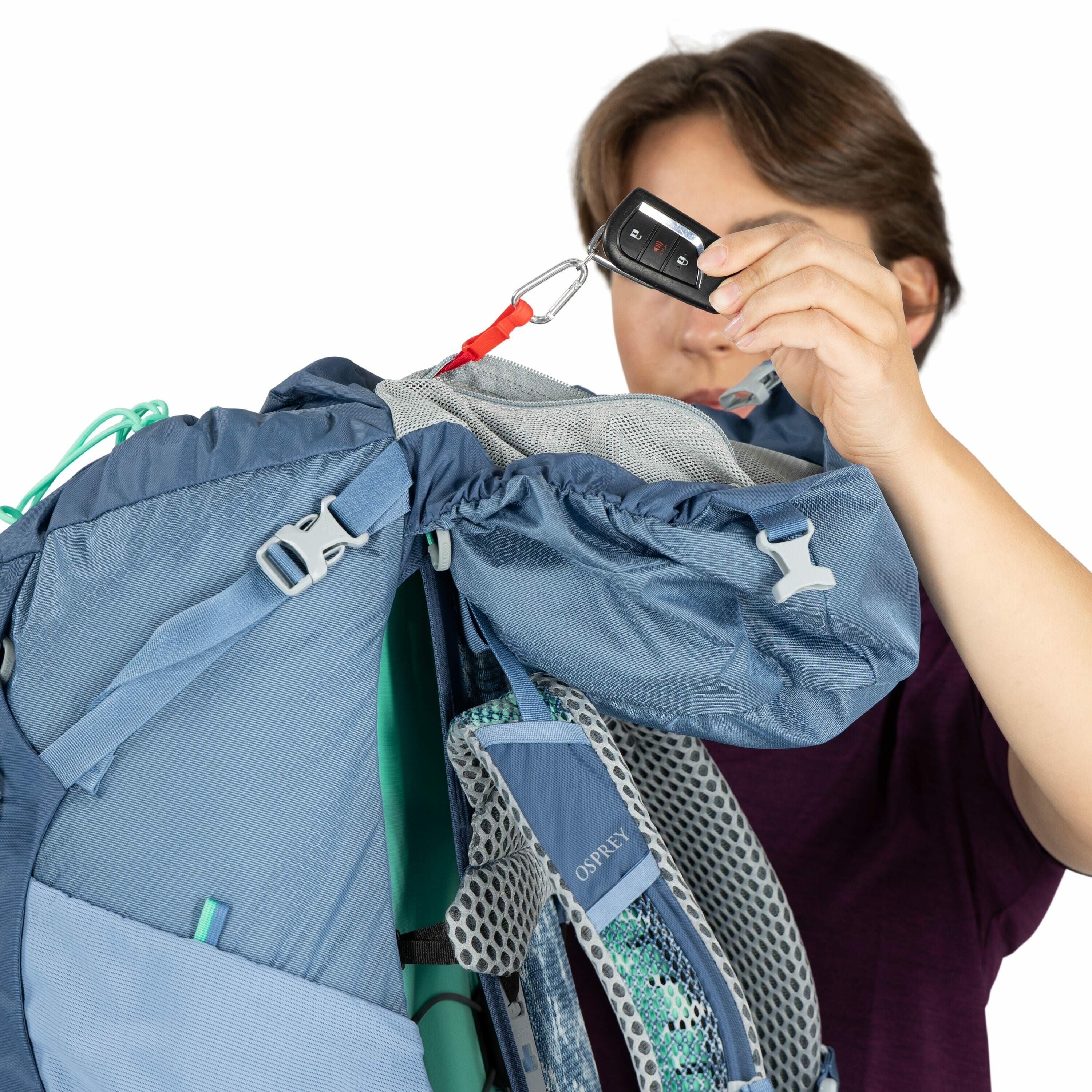 The image shows a person holding a blue Osprey backpack. The backpack appears to be large, suitable for hiking or backpacking, with adjustable straps and mesh padding for comfort and ventilation. The person is lifting the top flap of the backpack with a r