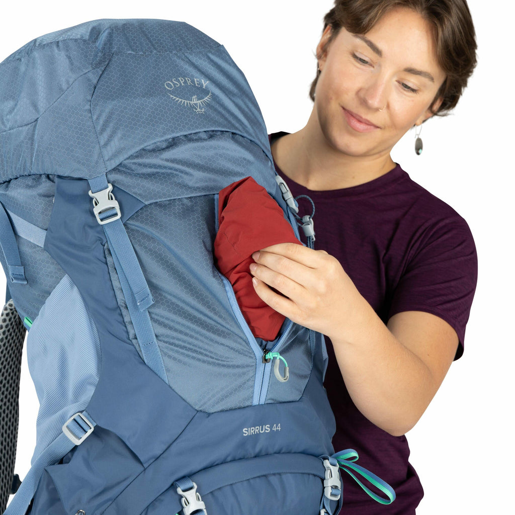 The image features a person holding a blue Osprey backpack with a visible "Osprey" logo and the model name "Sirrus 44" printed on the bag. The person is wearing a dark purple shirt and is inserting or removing a red item from a zippered pocket on the side