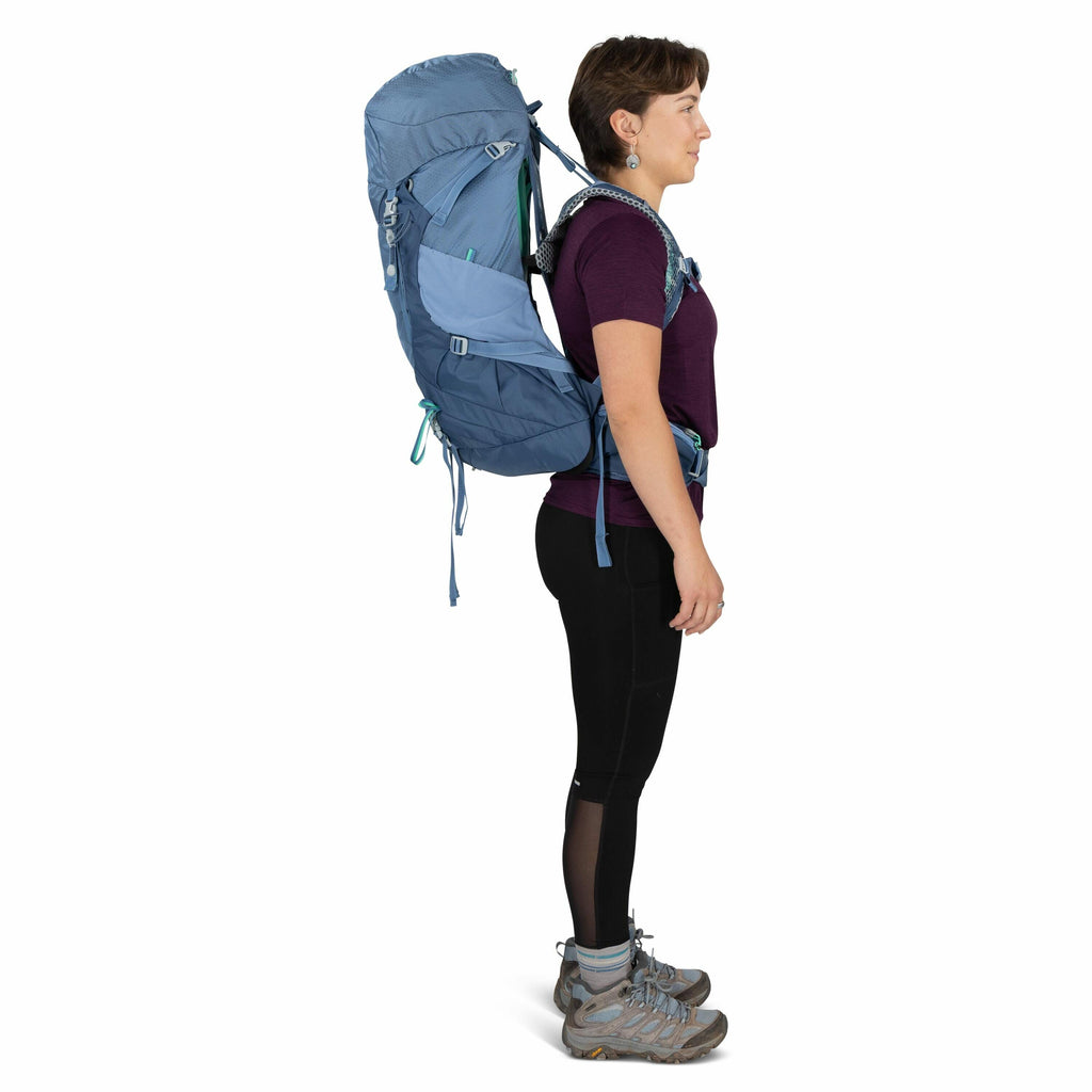 The image depicts a person standing in profile view, equipped for hiking or trekking. They are wearing a large, light blue backpack with multiple compartments and adjustable straps. The individual is dressed in a dark purple short-sleeve top, black leggin