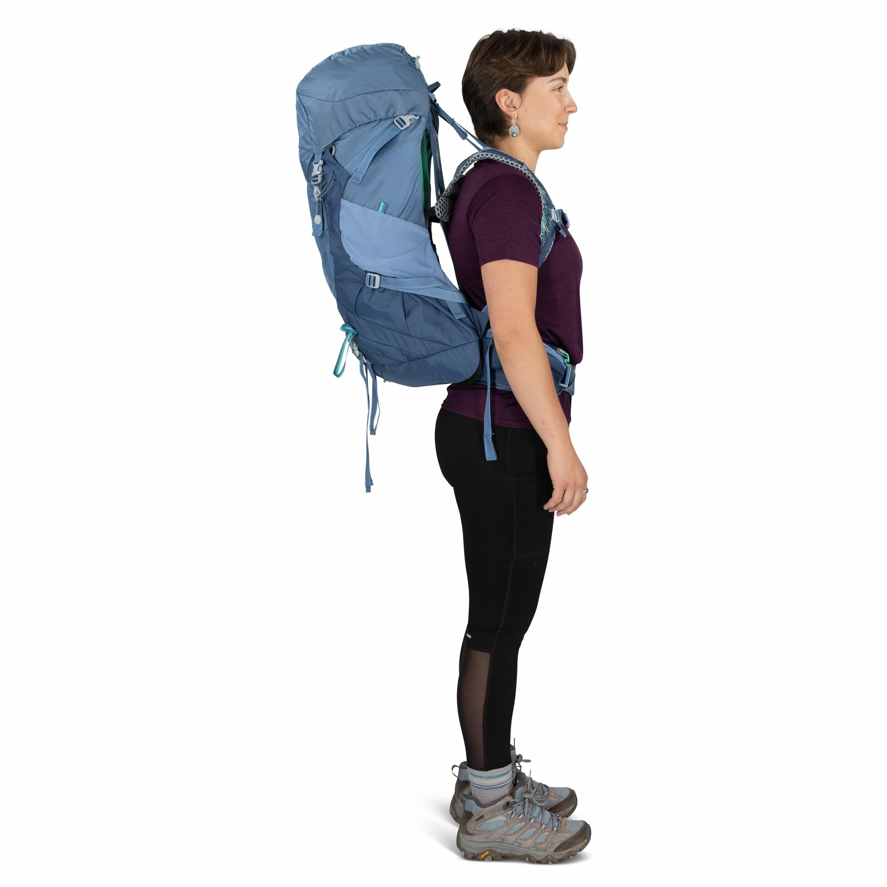 The image depicts a person standing in profile view, equipped for hiking or trekking. They are wearing a large, light blue backpack with multiple compartments and adjustable straps. The individual is dressed in a dark purple short-sleeve top, black leggin