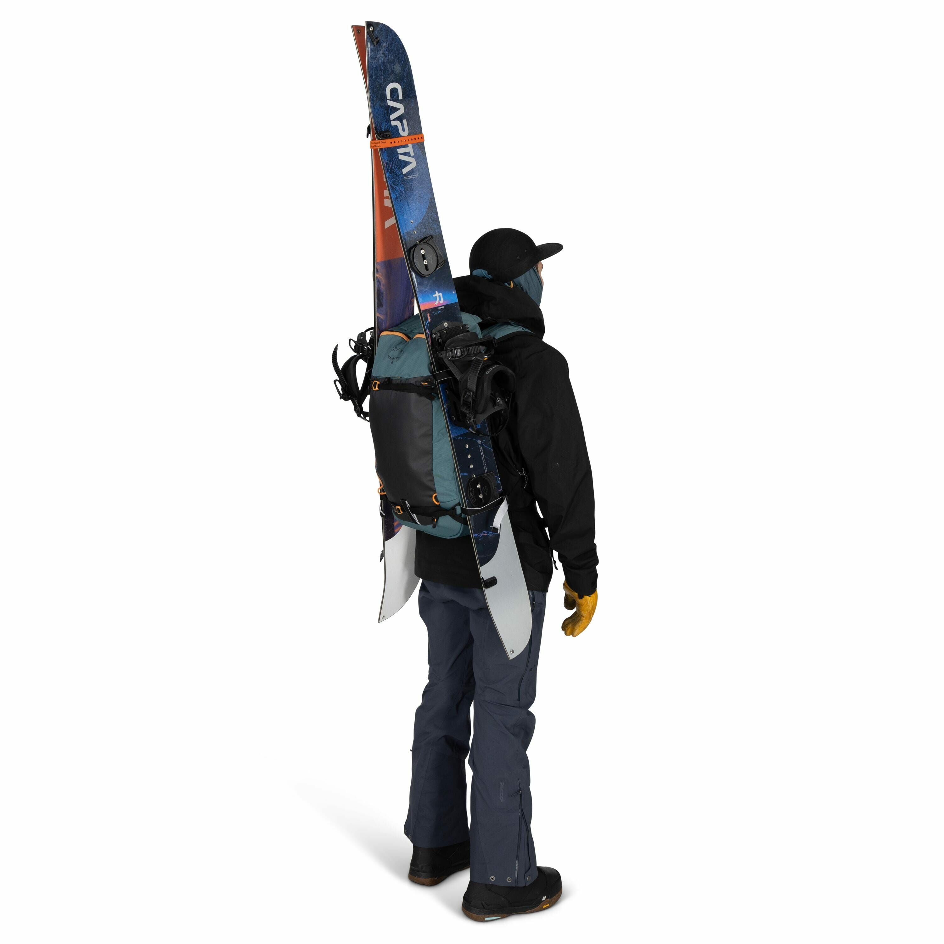 The image shows a person dressed in winter gear, viewed from the back. They are wearing a dark-colored jacket, a cap, and a pair of blue-gray ski pants. The person has a large backpack on their back with a pair of skis attached vertically. They are also w