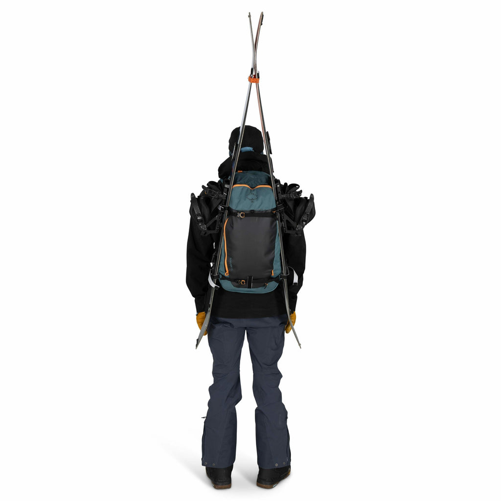 The image depicts a person viewed from the back, dressed in outdoor winter attire, likely prepared for skiing or snowboarding. They are wearing a black jacket, blue snow pants, and brown gloves, and carrying a large backpack. The backpack features a criss