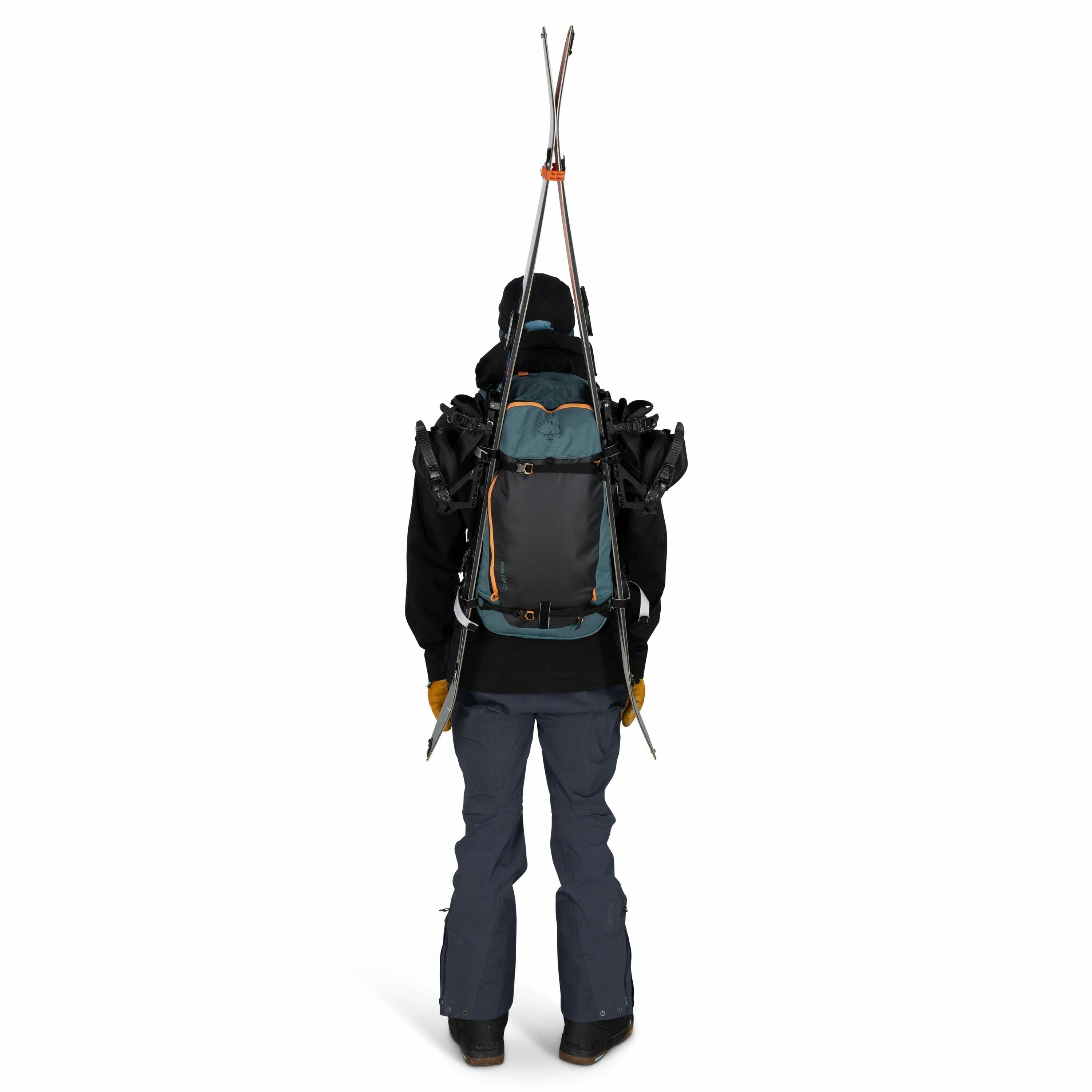 The image depicts a person viewed from the back, dressed in outdoor winter attire, likely prepared for skiing or snowboarding. They are wearing a black jacket, blue snow pants, and brown gloves, and carrying a large backpack. The backpack features a criss