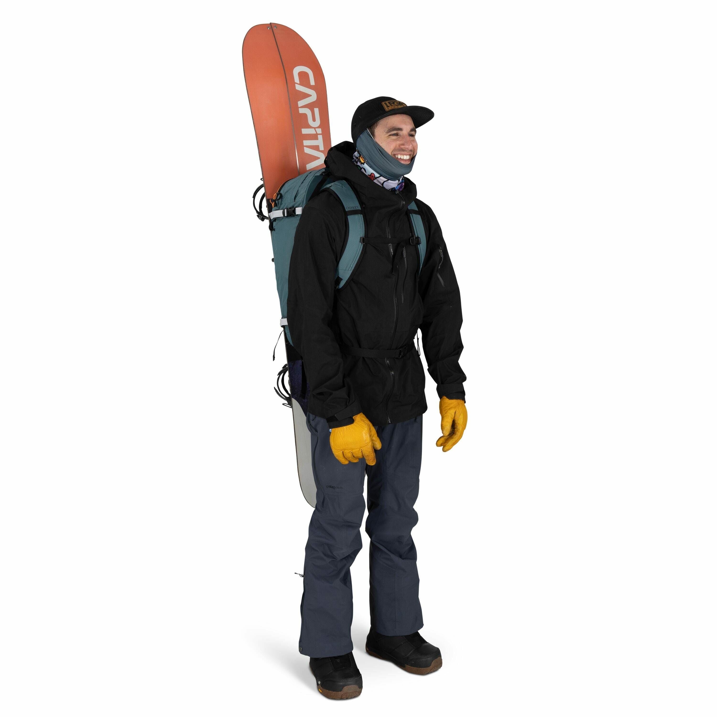 The image shows a person dressed in winter outdoor gear, likely prepared for snowboarding or skiing. They are wearing a black jacket, dark blue pants, and yellow gloves. A snowboard is secured vertically on their back with a backpack that has straps speci