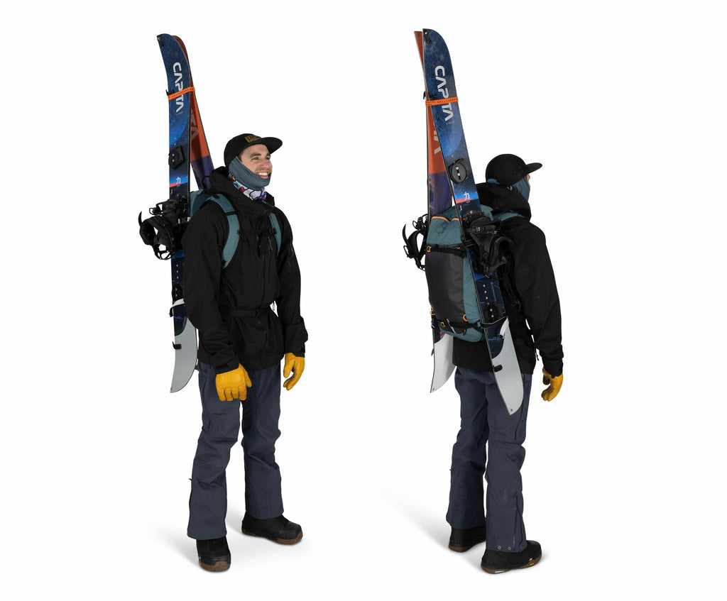 The image shows a person wearing outdoor winter gear suitable for skiing or snowboarding. They have two skis strapped to their backpack, positioned vertically and extending above their shoulders. The individual is dressed in a black jacket, dark pants, an