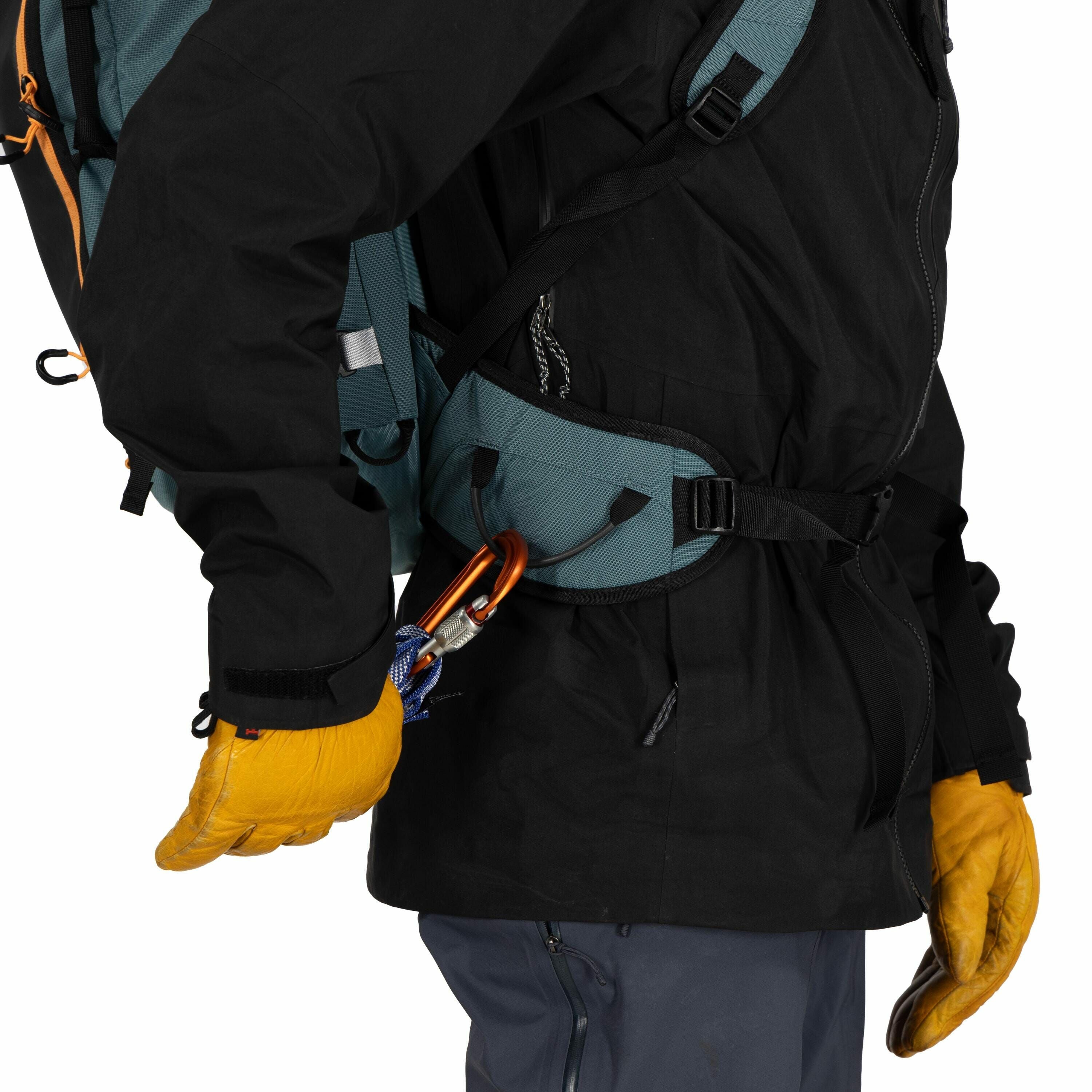 The image shows a person dressed in outdoor gear, suitable for hiking or climbing. They are wearing a black jacket, gray pants, and yellow gloves, and are seen from the side. The individual is carrying a large backpack with a turquoise and orange color sc
