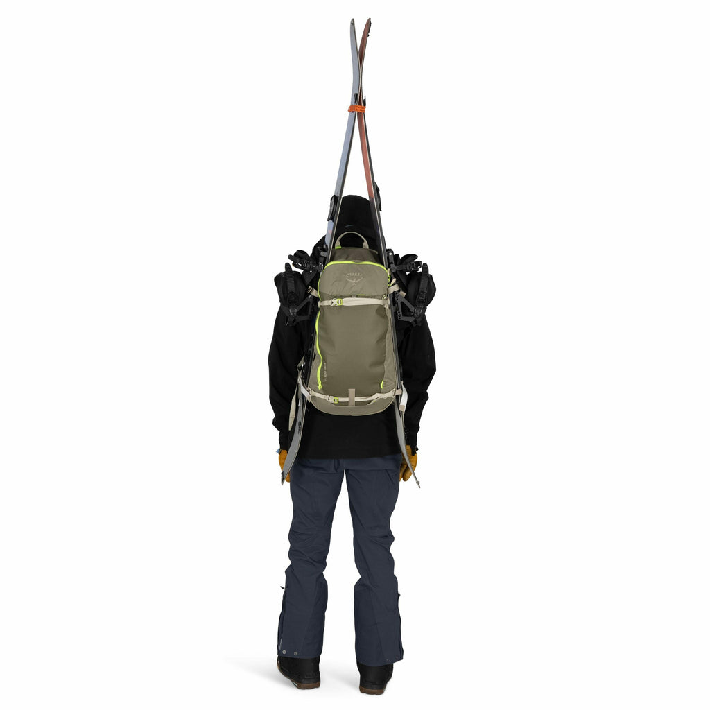 The image shows a person dressed in outdoor winter clothing, seen from the back. They are wearing a dark jacket with a hood, and blue snow pants, along with black boots. The person is carrying an olive green backpack with lime green accents, which has mul