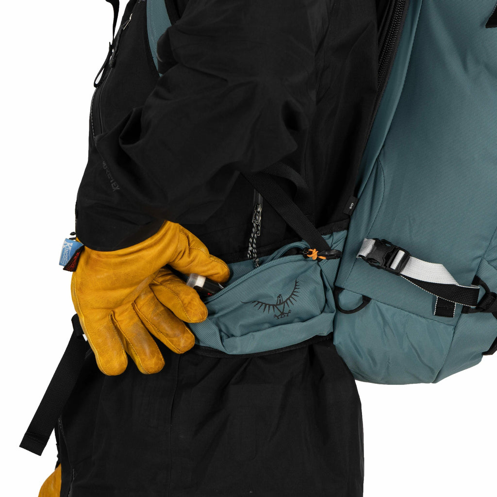 The image shows a person wearing a black jacket and carrying a teal-colored backpack with a visible hip belt pouch. The person is reaching into the pouch with their right hand, which is wearing a bright yellow glove, indicating they might be preparing for