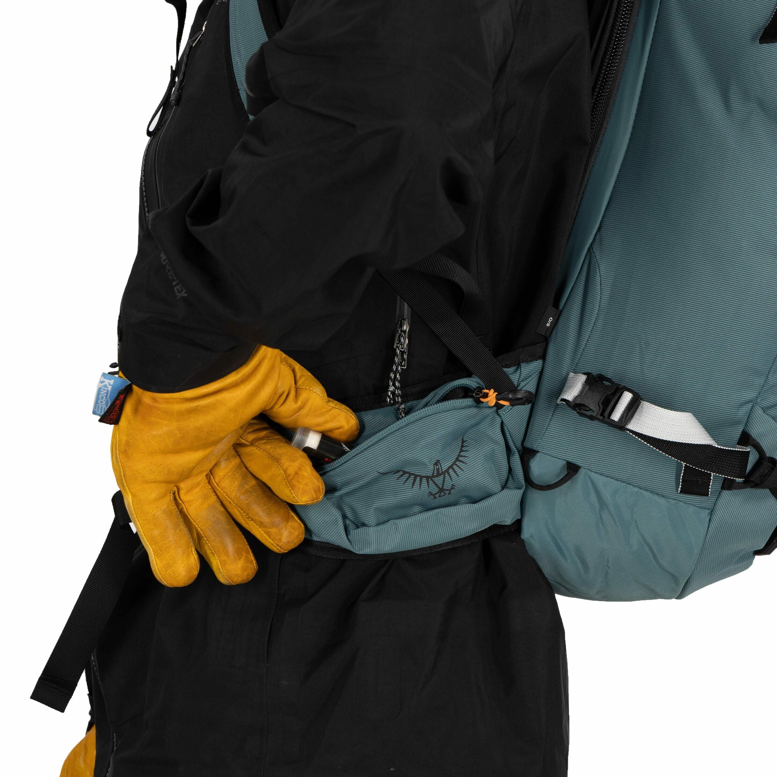 The image shows a person wearing a black jacket and carrying a teal-colored backpack with a visible hip belt pouch. The person is reaching into the pouch with their right hand, which is wearing a bright yellow glove, indicating they might be preparing for